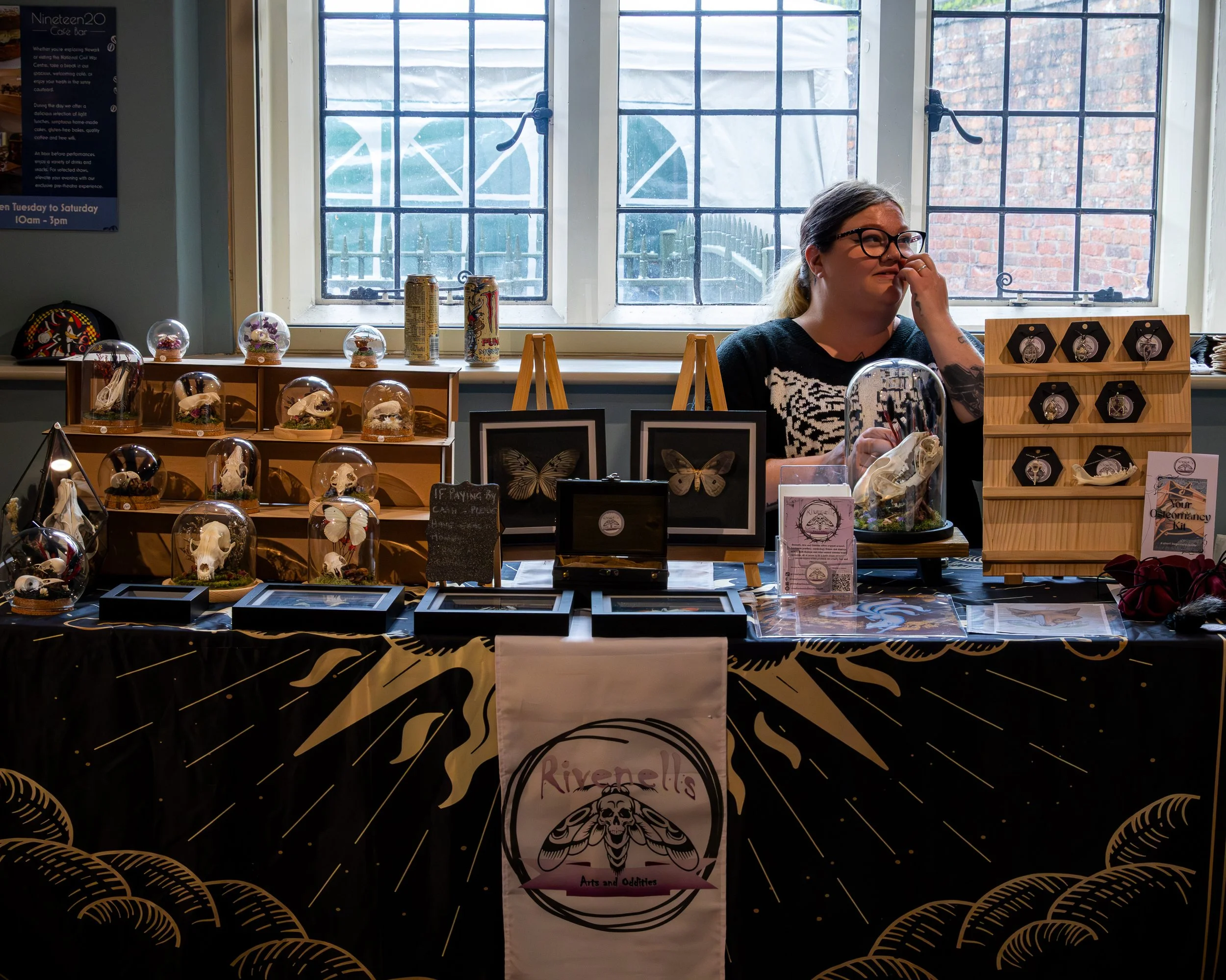 A woman wearing glasses behind a display table with various art and collectible items in a shop or market, with three large windows behind her.