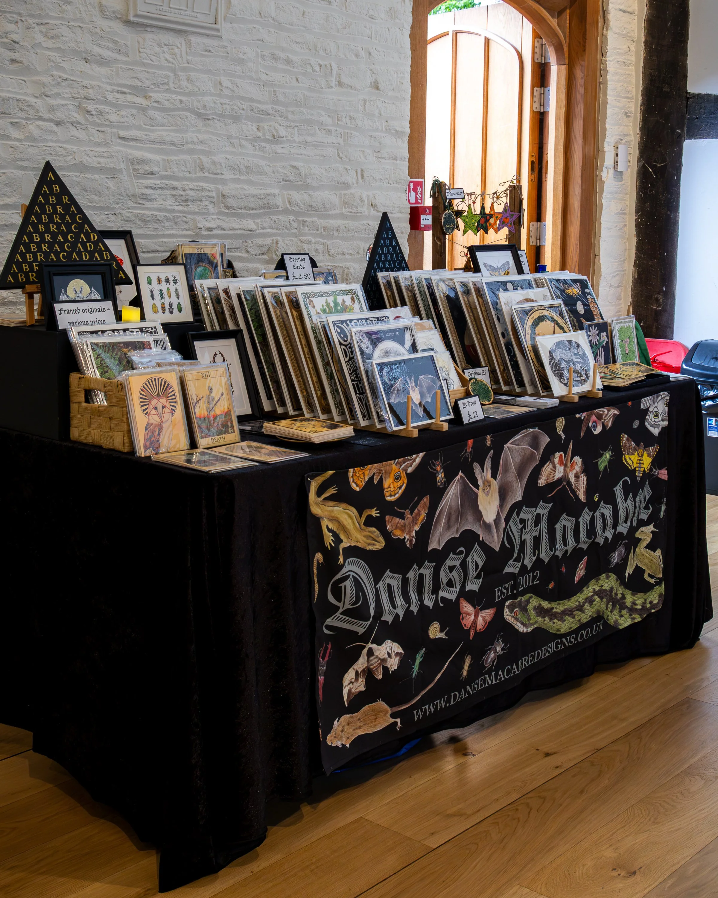 A display table with various framed artwork, greeting cards, and crafts at an indoor market or store. The table is draped with a black cloth featuring images of moths, butterflies, and reptiles, and has signs indicating prices and product description