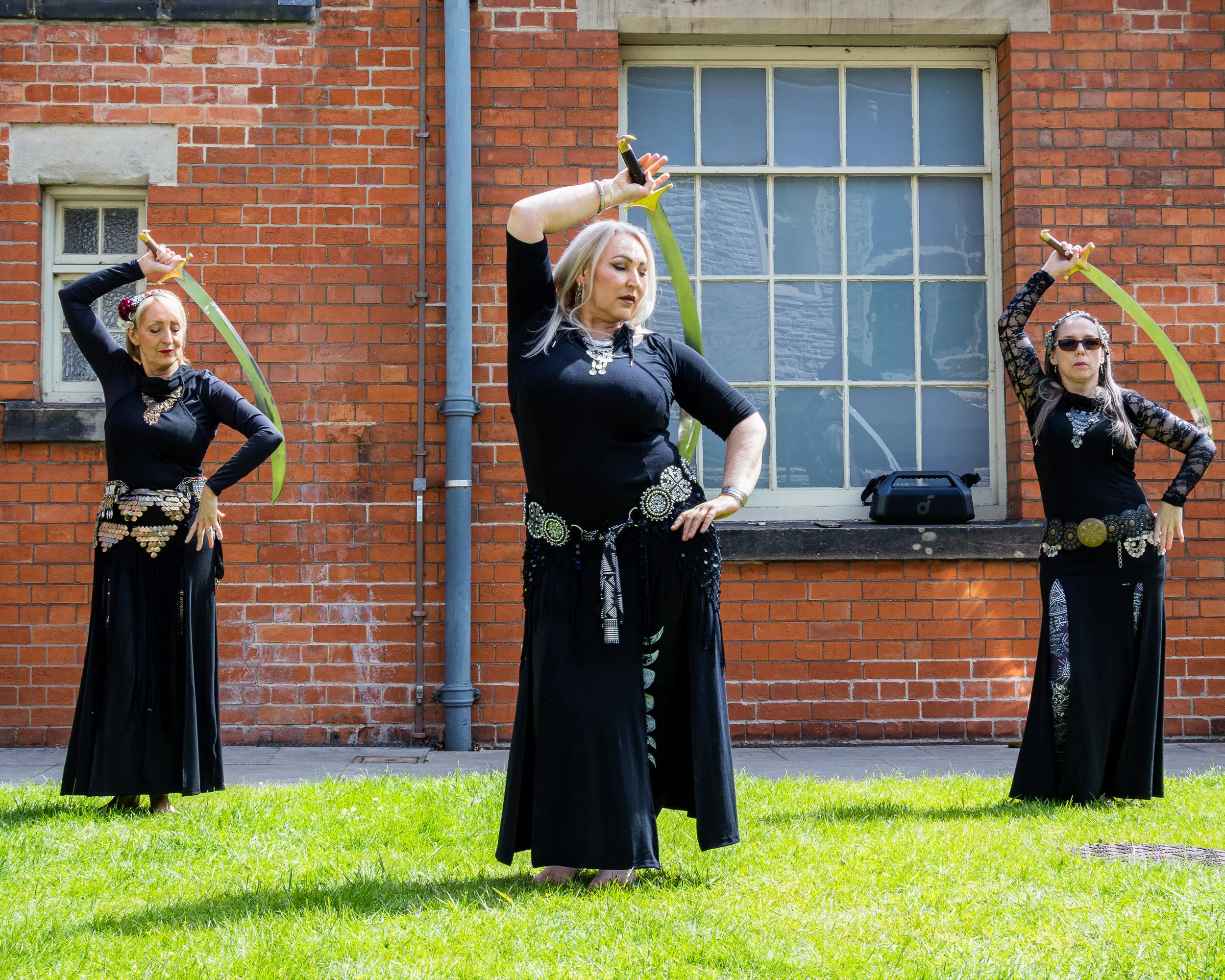 Three women dressed in black with artistic accessories performing a dance outdoors on a grassy area in front of a brick building with large windows.