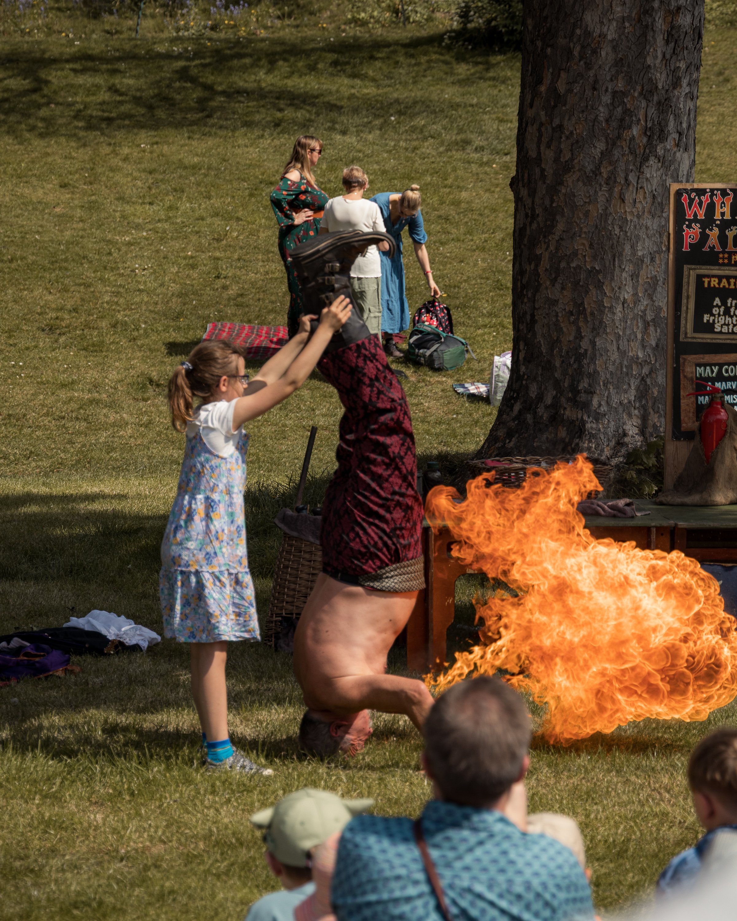 Group of children and women watching and participating in a fire-bighting demonstration outdoors on grass, with some children sitting and raising their hands, and a fire in a controlled fire pit.