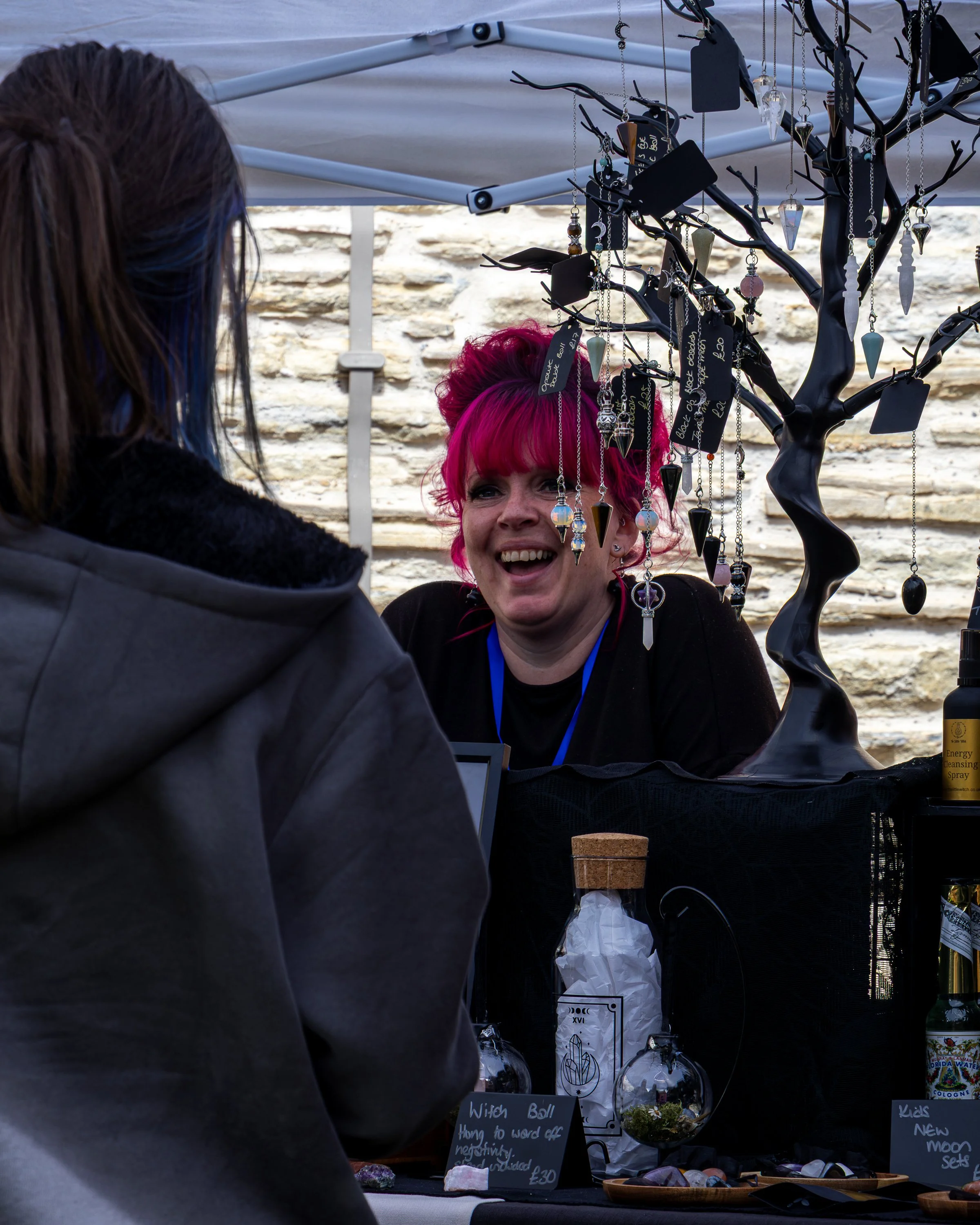 A woman with bright pink hair is smiling at a table of jewelry and crystals while talking to another person under a canopy. The table displays hanging necklaces, some with pendants, and various rocks or crystals in bowls. Background shows a stone wal