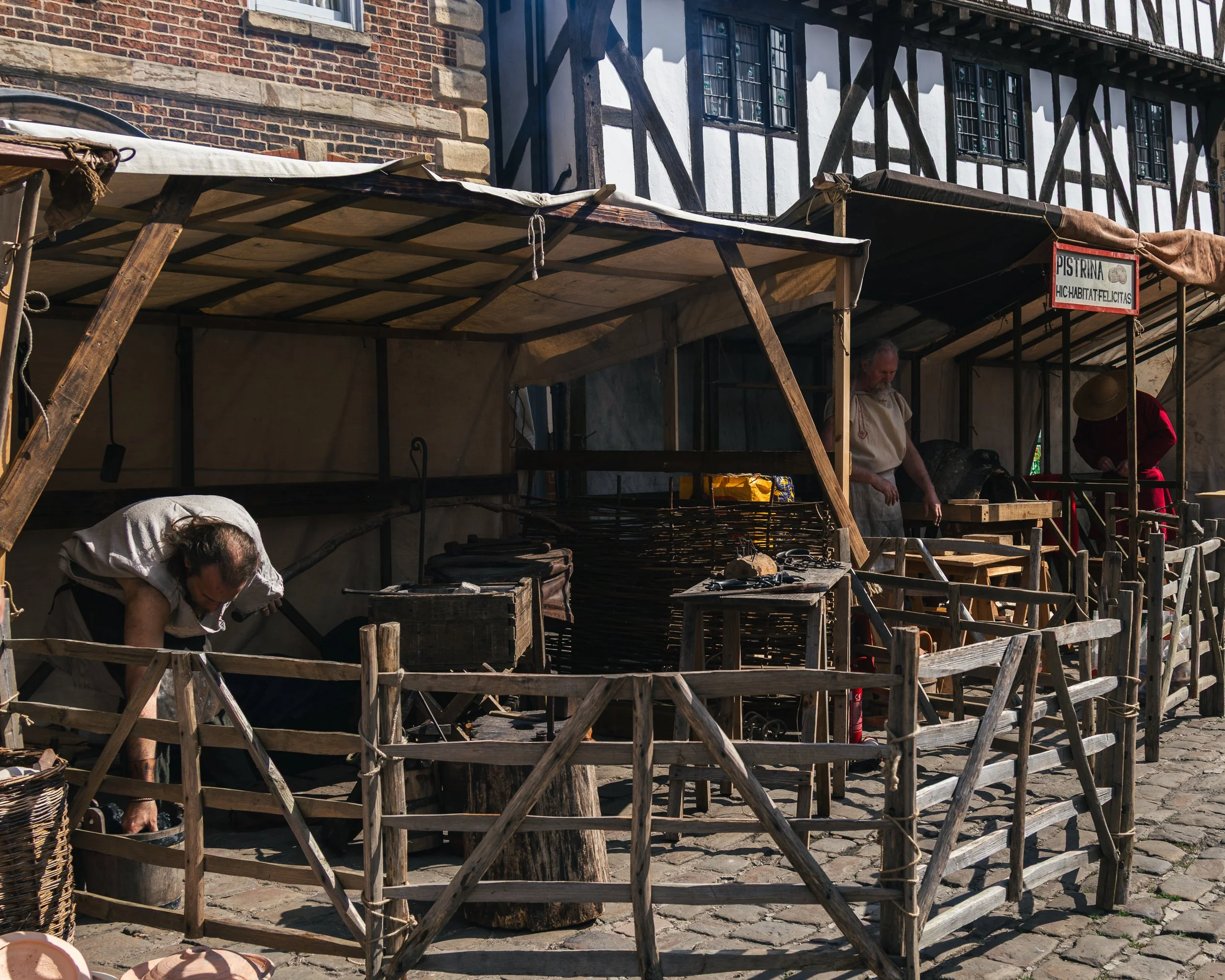 A historical market stall setup with wooden scaffolding and fabric covering, two people visible, one bending over and the other standing, in front of a half-timbered building with windows with stained glass.