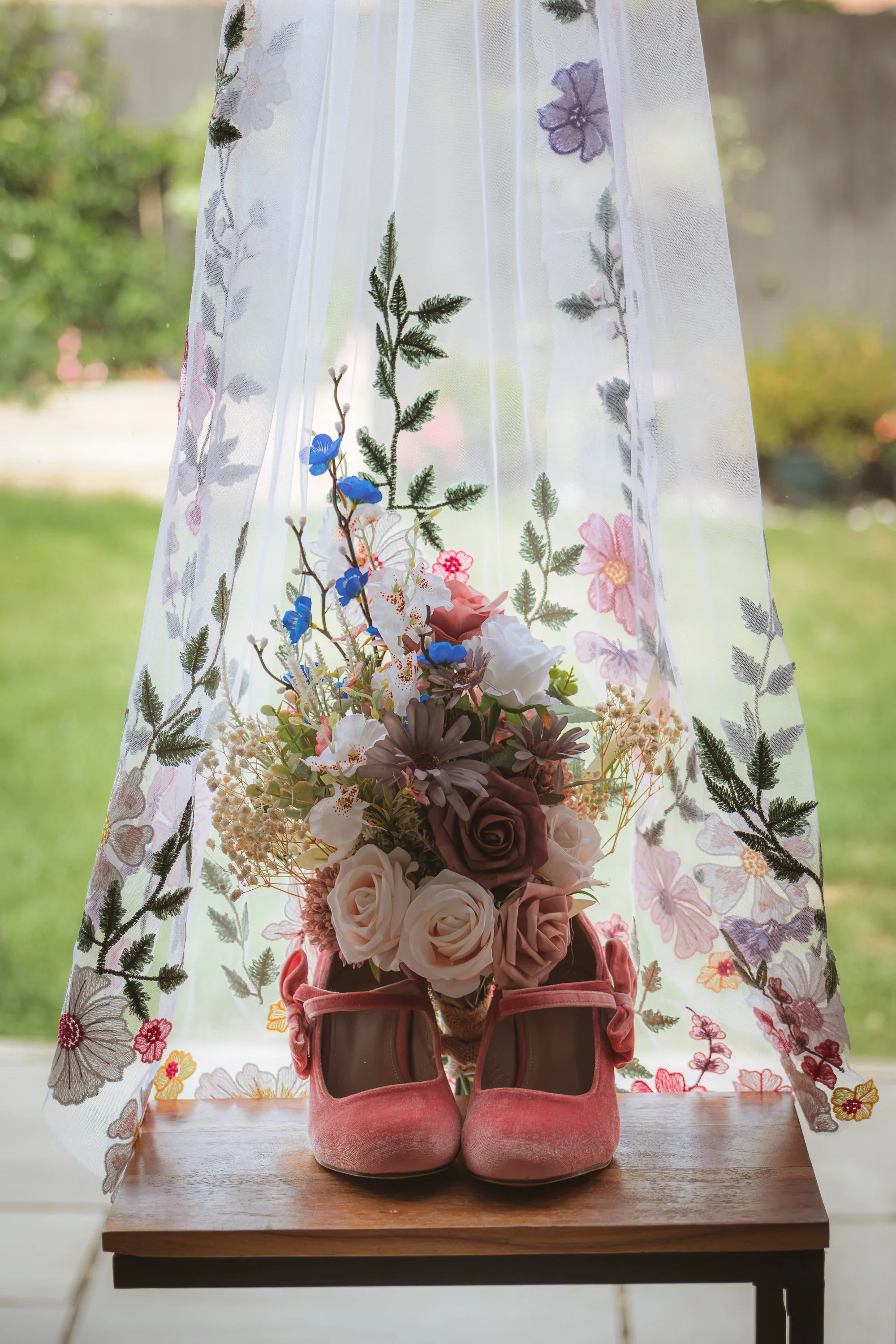 A pair of pink shoes with heels, a floral bouquet, and a botanical decoration sit on a wooden table in front of a sheer curtain with floral embroidery. Outside view is blurred in the background.
