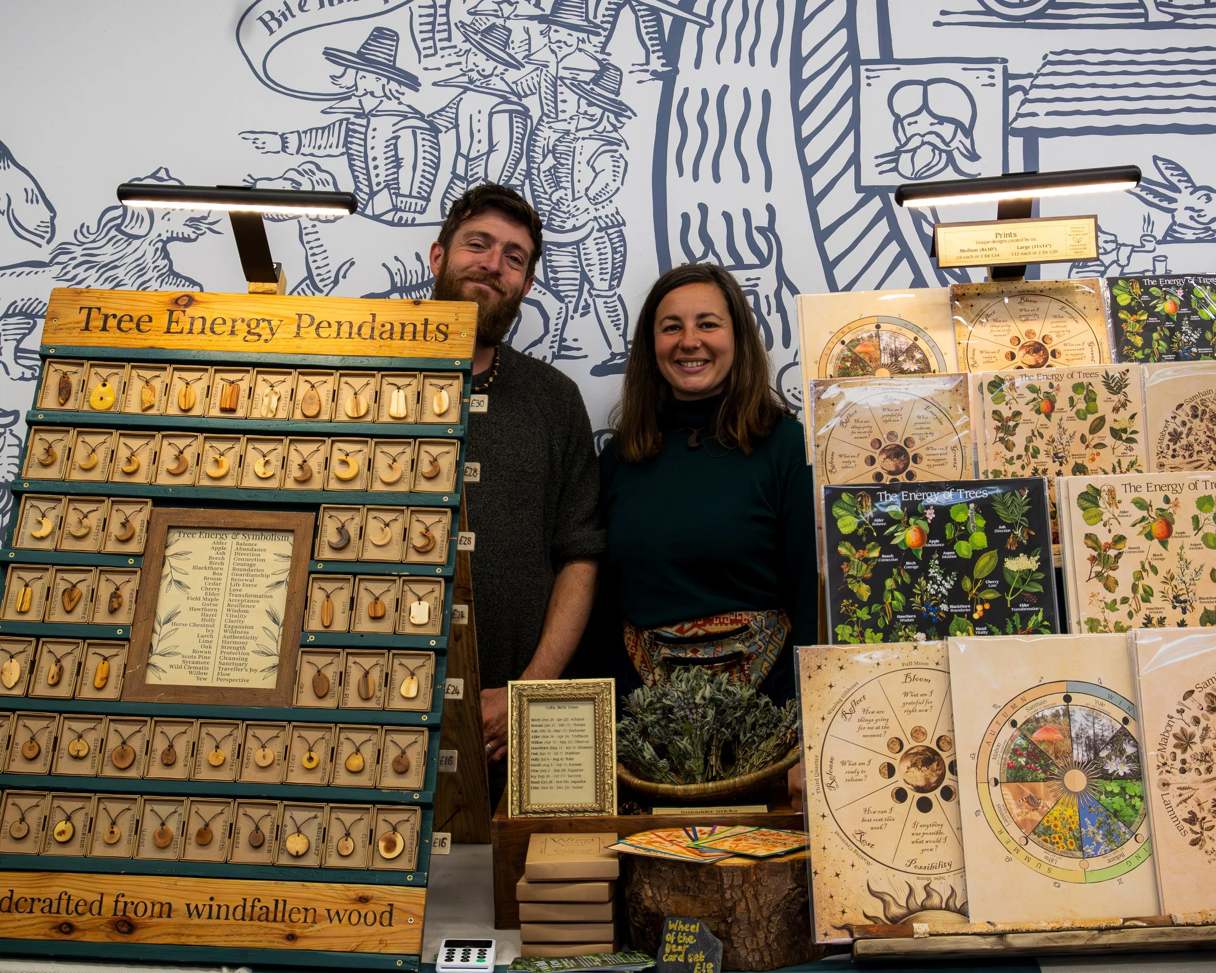 Two smiling women standing behind a table displaying tree-themed jewelry and educational materials at an indoor event. The jewelry includes pendants shaped like moons and leaves, labeled as 'Tree Energy Pendants.' Behind them, a colorful wall mural d