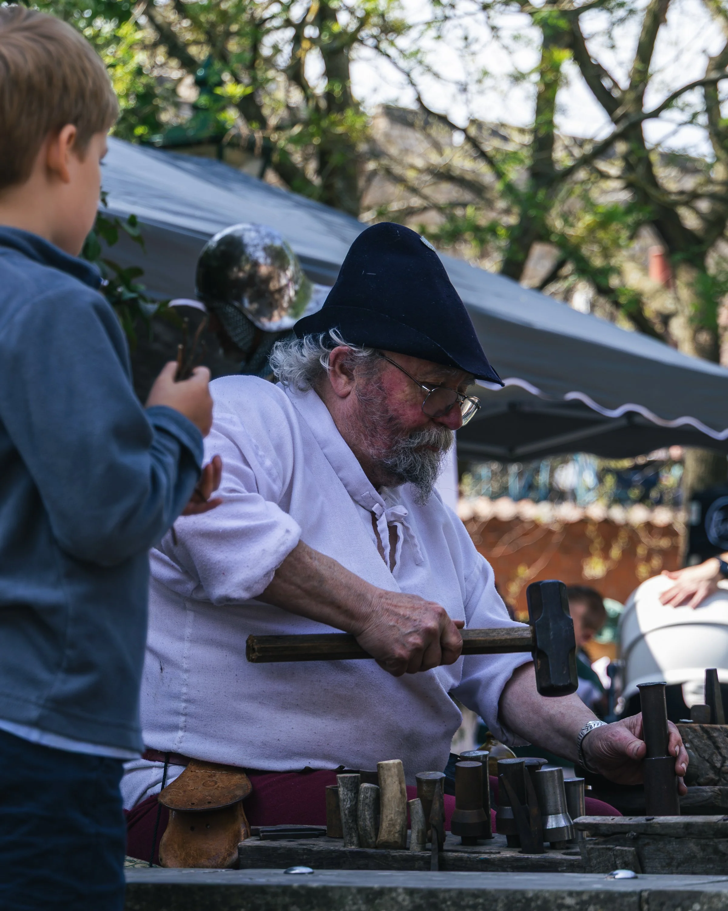 A man with a beard and glasses wearing a black hat and white shirt working with tools on a wooden surface outdoors, with a young boy observing.