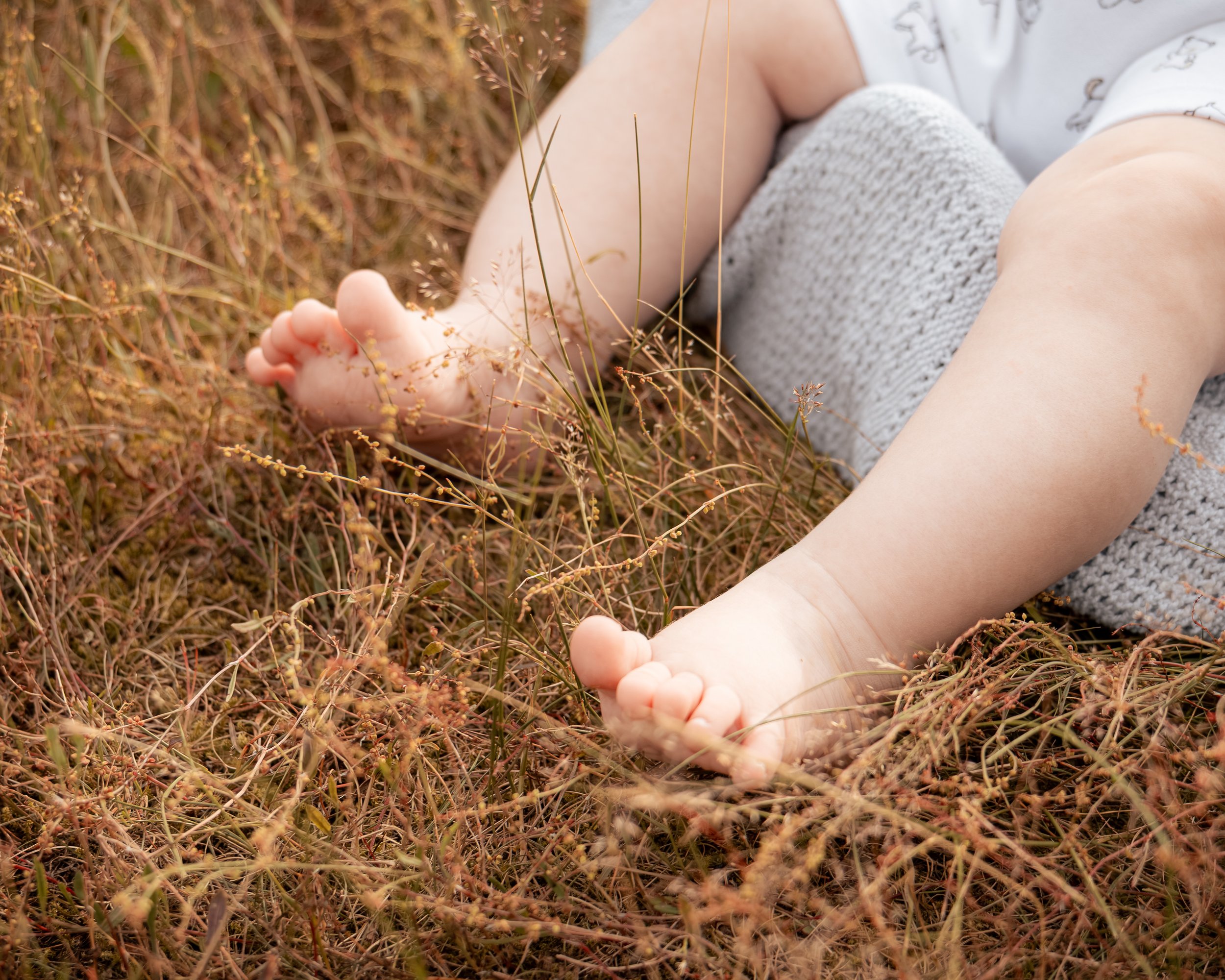 A child sitting on the ground among dry grass and weeds, wearing a light gray knitted sleeve and a white T-shirt with a cartoon print, reaching out with their hand.