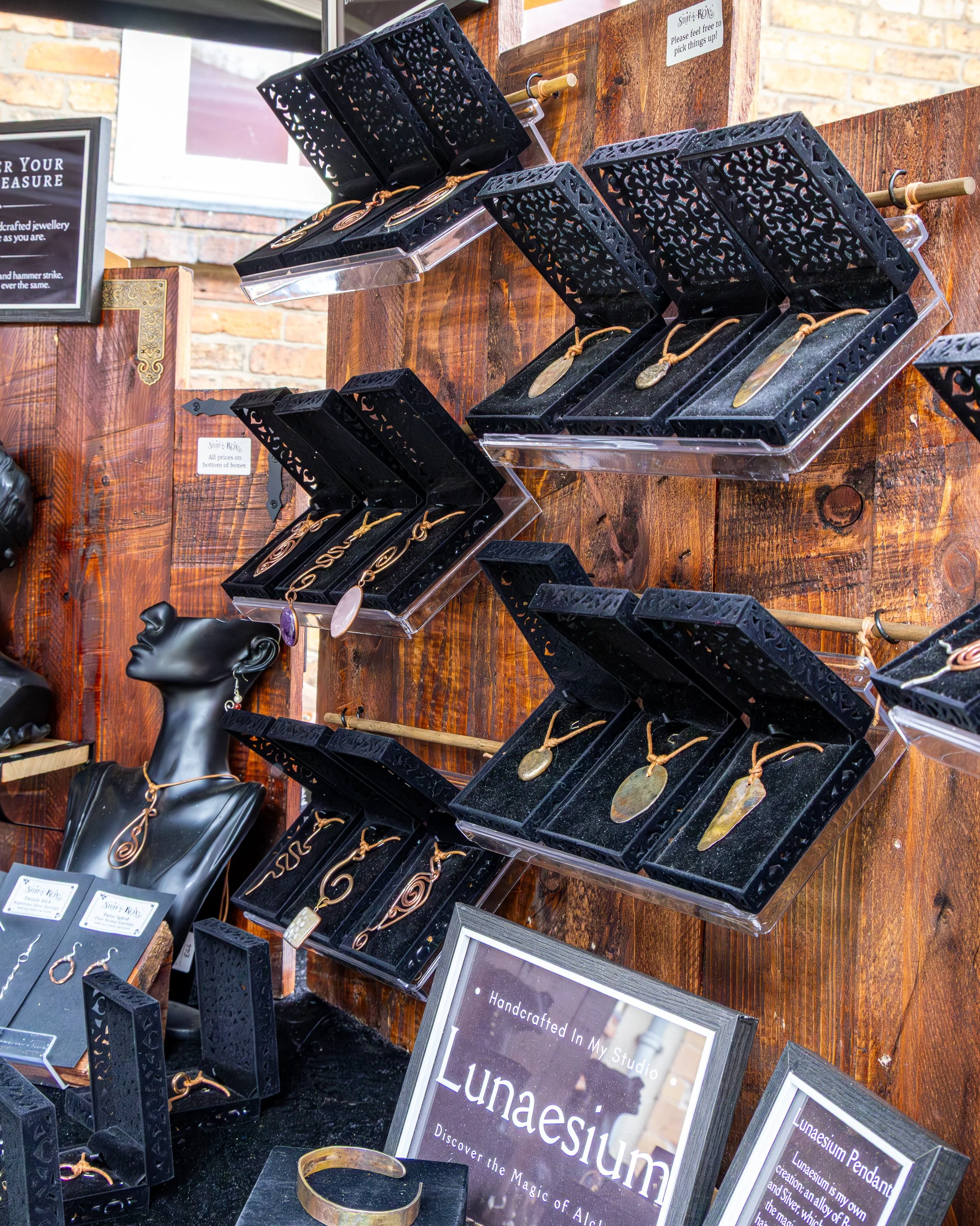 Display of handcrafted jewelry, including necklaces and bracelets, on black bust mannequins and black boxes with jewelry inside. The jewelry features gold and bronze tones with decorative designs. There are signs and frames with information about Lun