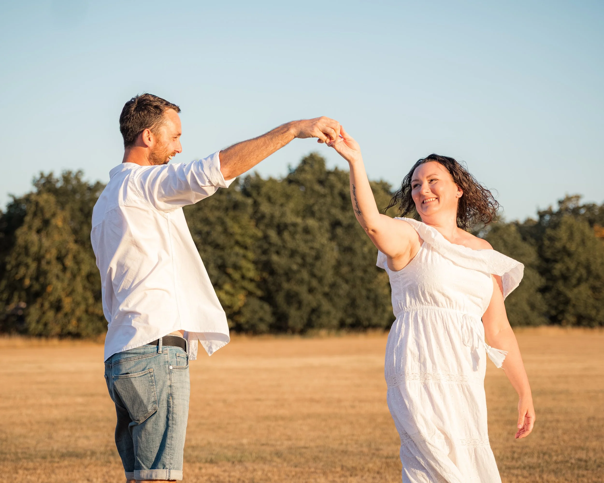 A couple dancing outdoors during sunset with a field and trees in the background, holding hands and smiling.