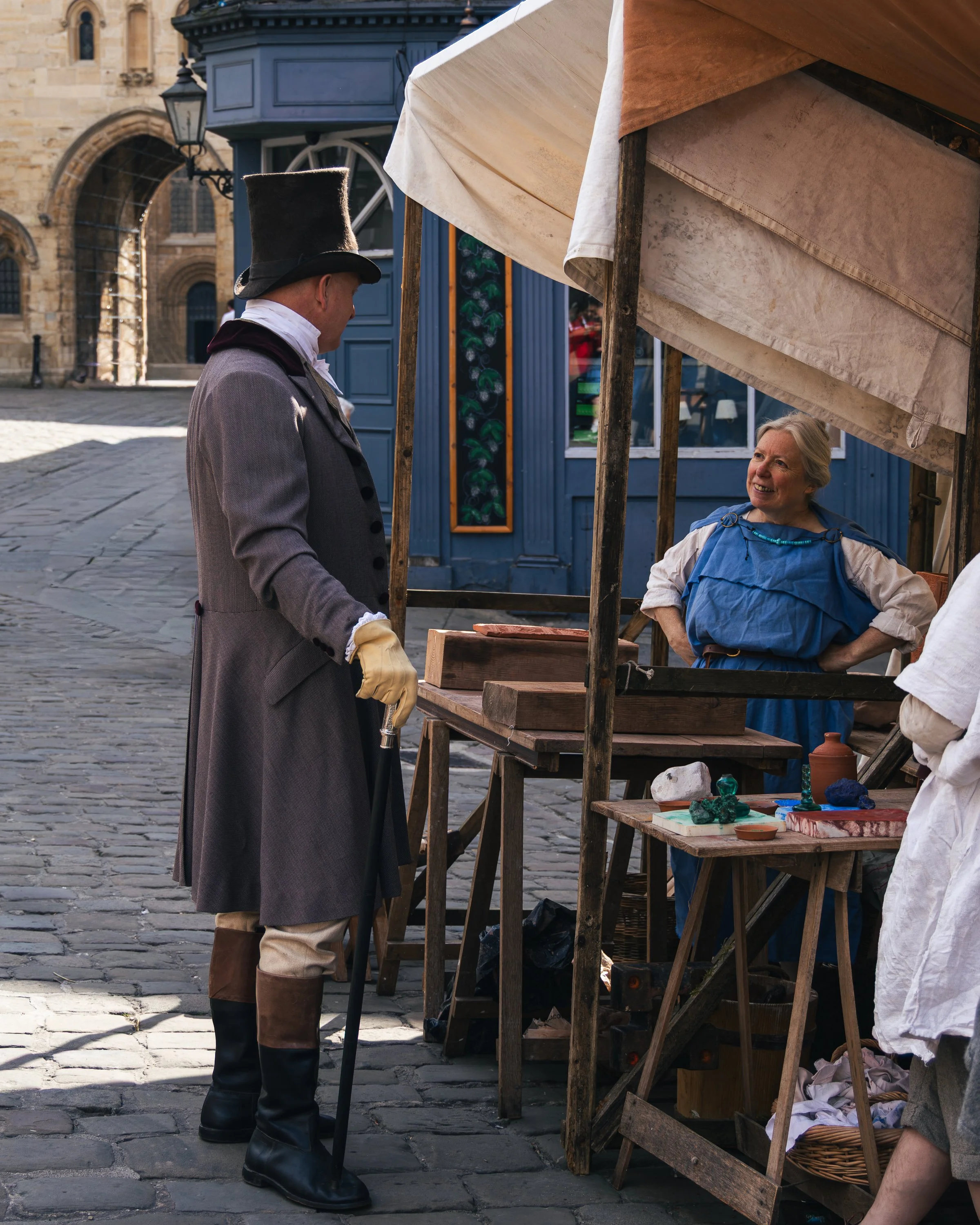 A man dressed in historical clothing, including a gray coat, beige trousers, and tall black riding boots, is standing at a market stall. He is wearing a tall black top hat and appears to be talking to an older woman behind the stall. The woman, with 