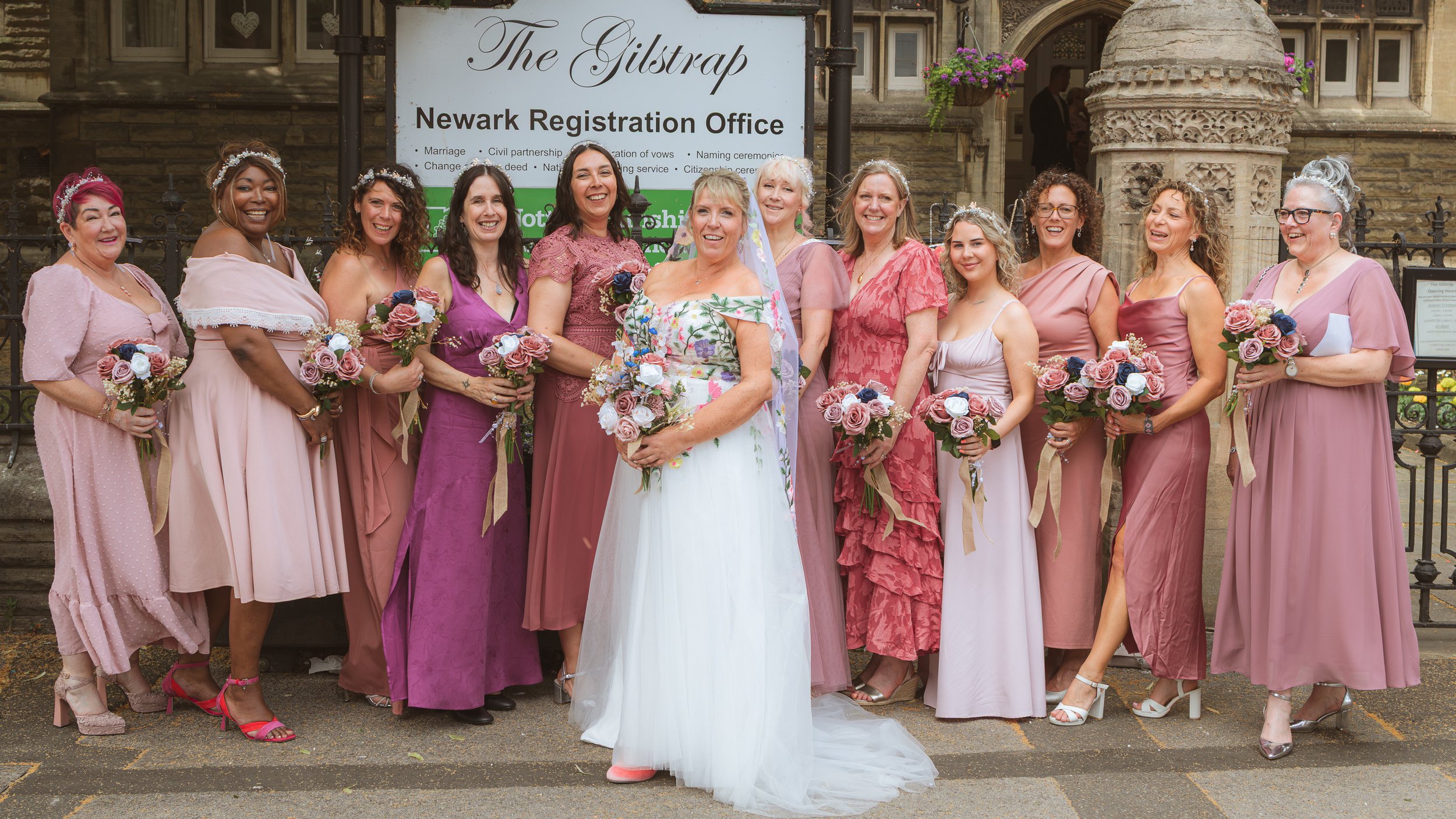 A group of women dressed in pink and purple dresses, holding bouquets, standing in front of a building with a sign that reads 'The Gilstrap, Newark Registration Office'.