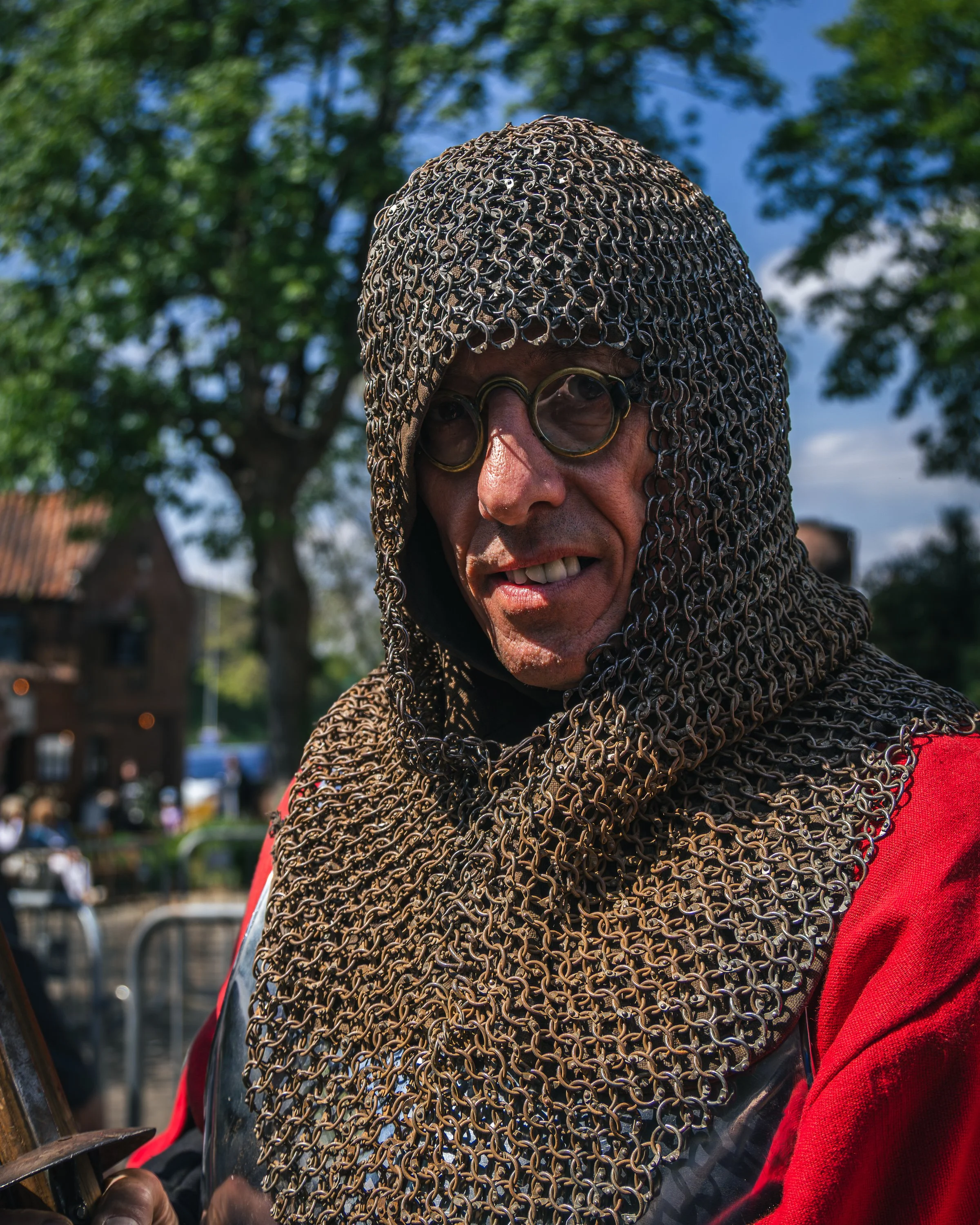 A person wearing medieval-style chainmail armor and round glasses, outdoors with trees and a blue sky in the background.