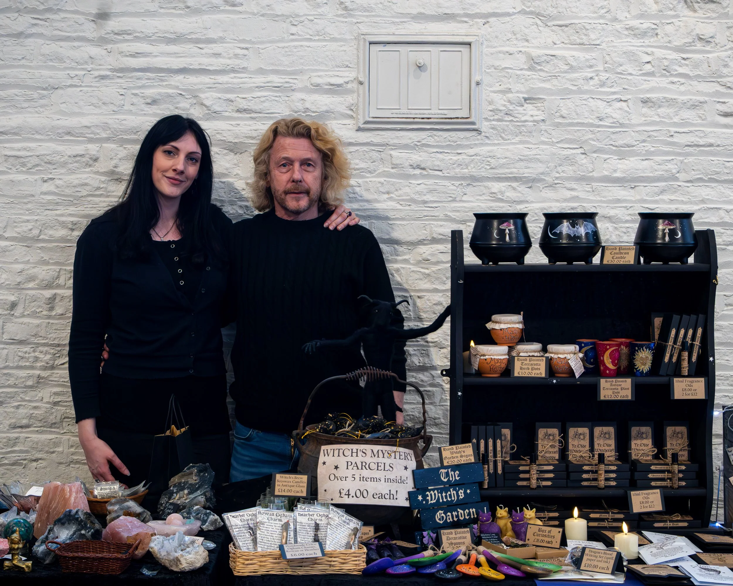 Two women standing behind a table at a market stall selling witch-themed items. The table displays various stones, candles, and decorative objects. Signage advertises witch's mystery parcels, hand painted cauldrons, and other mystical items. The back
