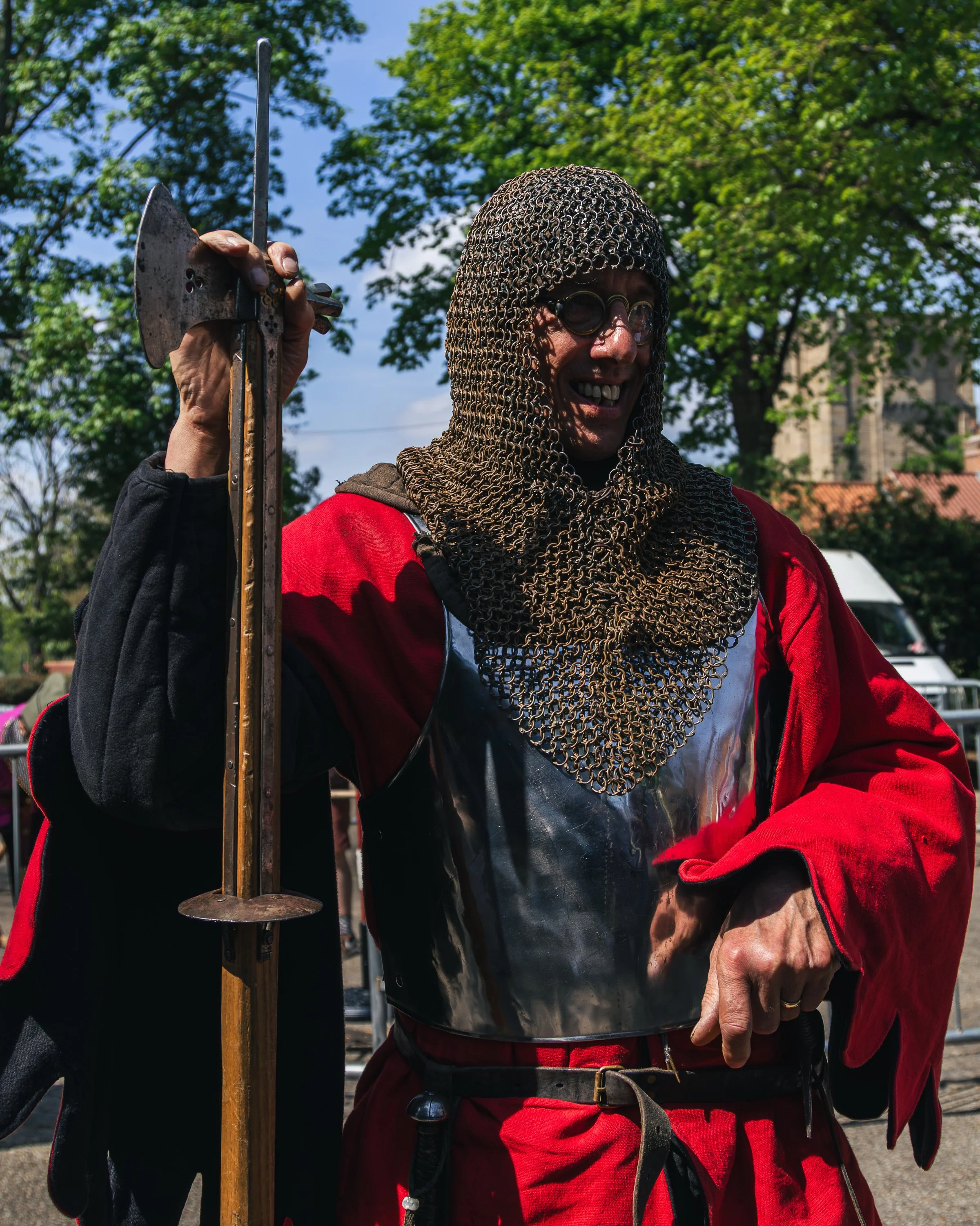 Person smiling wearing chainmail hood and glasses, holding a metal sword, outdoors with trees and buildings in background.