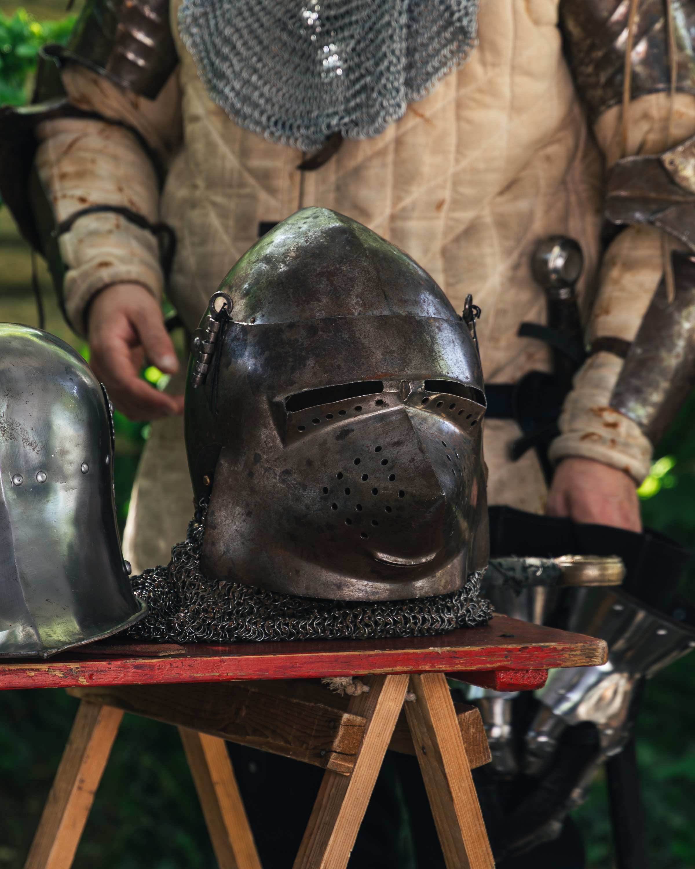 A person dressed in medieval armor, including a chainmail hood and a metal helmet, is displayed on a wooden table outdoors. The background is blurred with greenery.