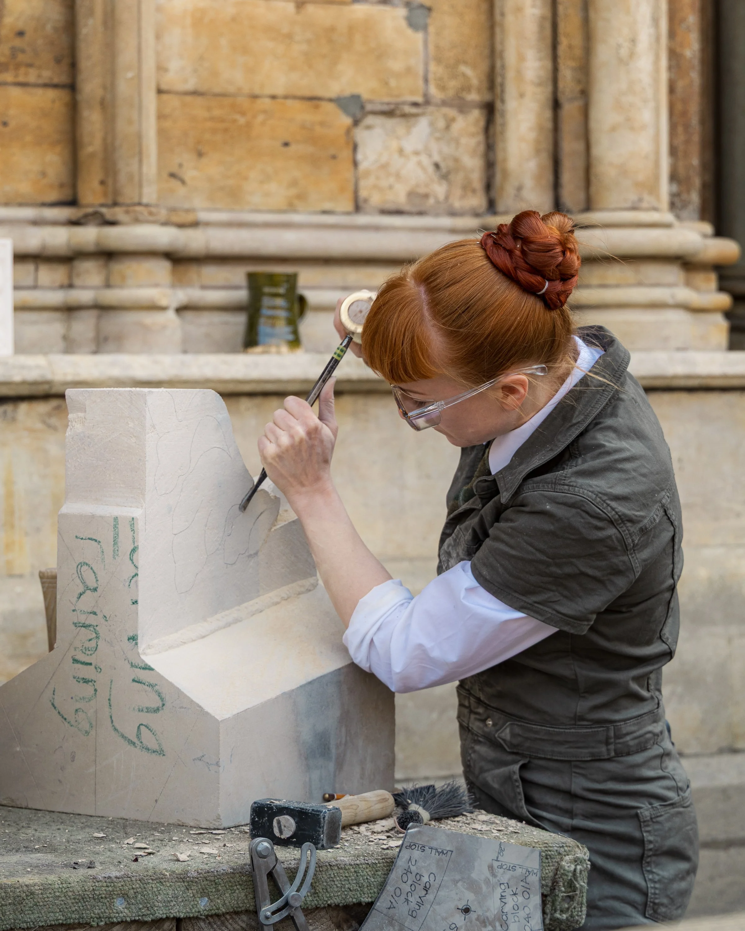 A woman with red hair in a bun, wearing gloves and safety glasses, carves stone in a workshop. She is working on a large piece of stone, which is supported on a sturdy surface with tools and safety equipment around her.