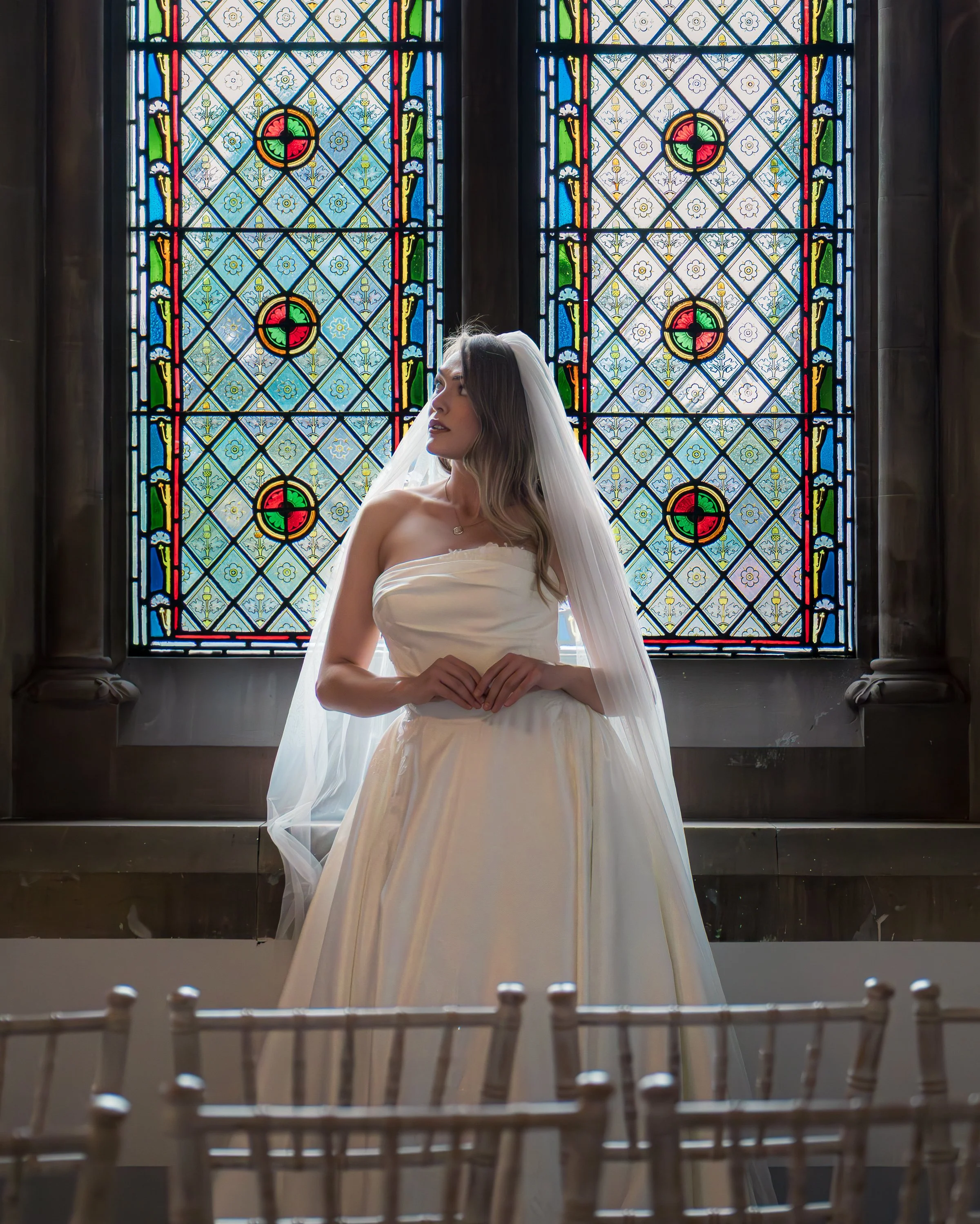A woman in a wedding dress and veil standing in front of stained glass windows, with wooden chairs in the foreground. The Renaissance at Kelham Hall