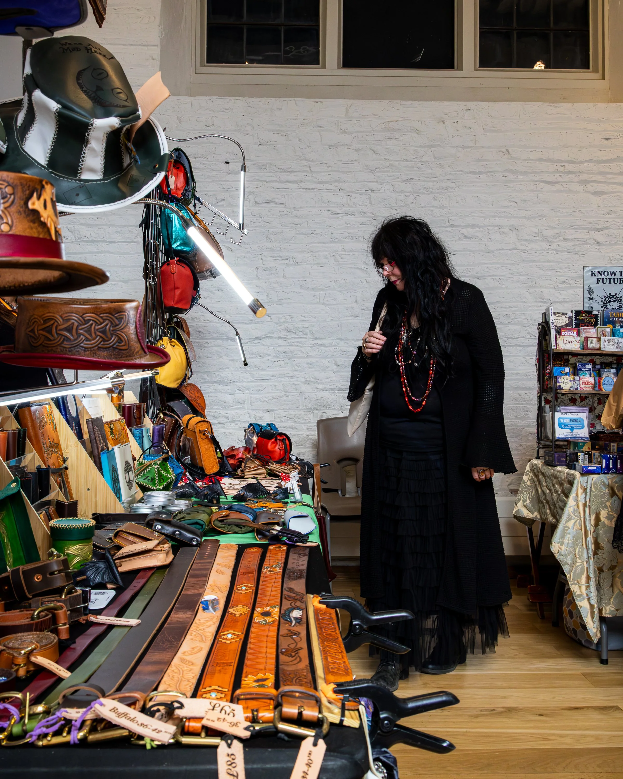 A woman with long dark hair and glasses browsing a table of leather belts, wallets, and accessories at an indoor market. The table displays various colorful leather products, and a small display of books is visible in the background against a white b
