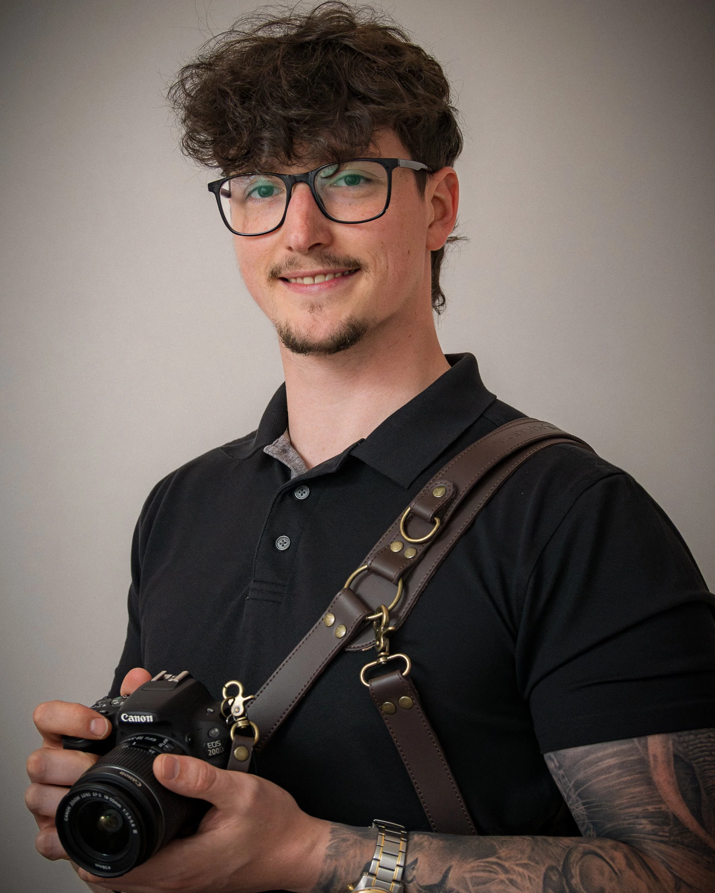 Close-up of a young man with glasses smiling and holding a Canon DSLR camera, wearing a black shirt and a brown leather strap bag, with visible tattoos on his forearm, against a plain background.