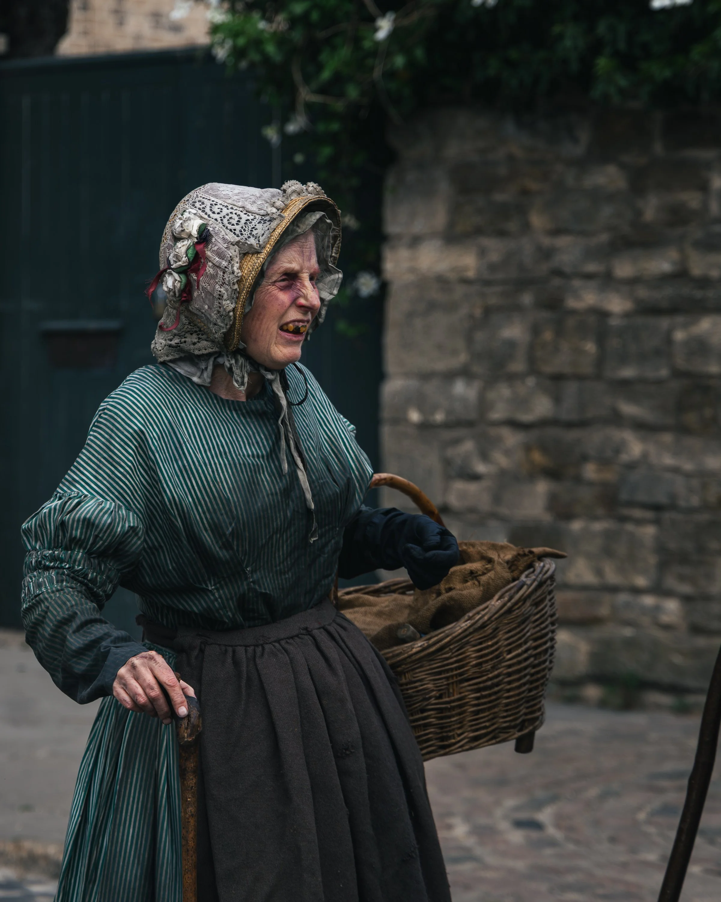 Old woman wearing a traditional bonnet, striped dress, and dark apron, walking outdoors with a basket of items, holding a walking stick.