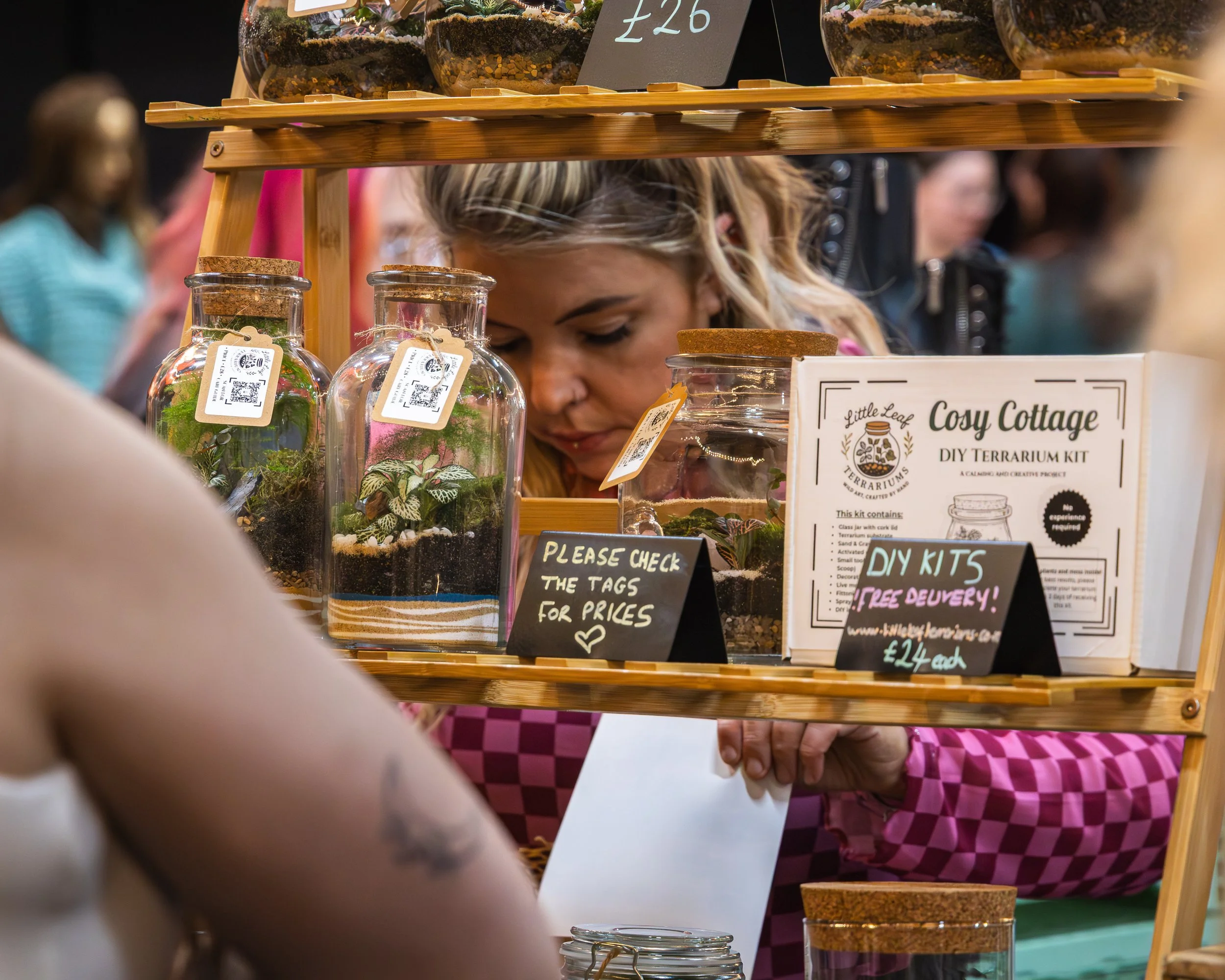Display of terrarium kits with glass jars containing plants and soil, at a market stall with signs and a crowd in the background.
