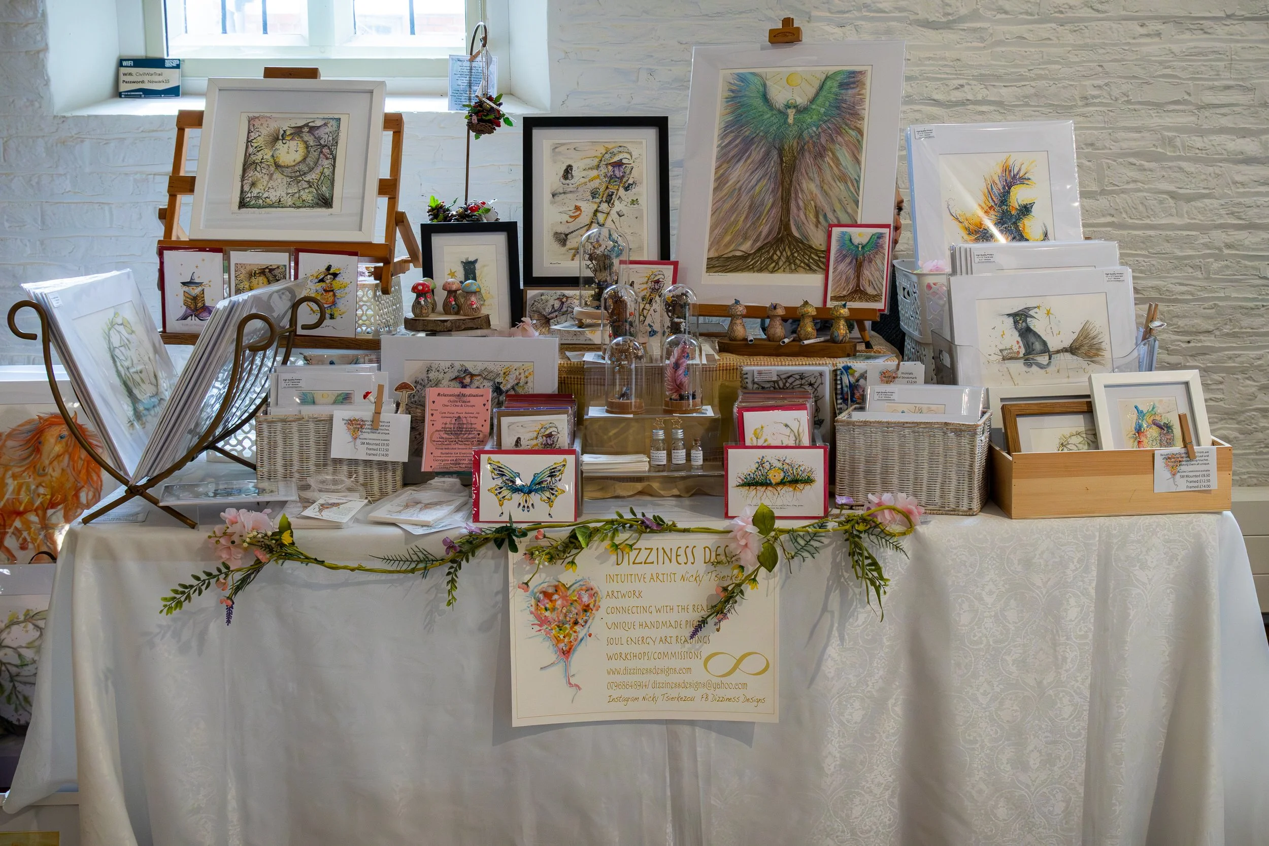 Art display table featuring framed illustrations of mythical creatures and angels, with decorative mushrooms and small items, set against a white brick wall.