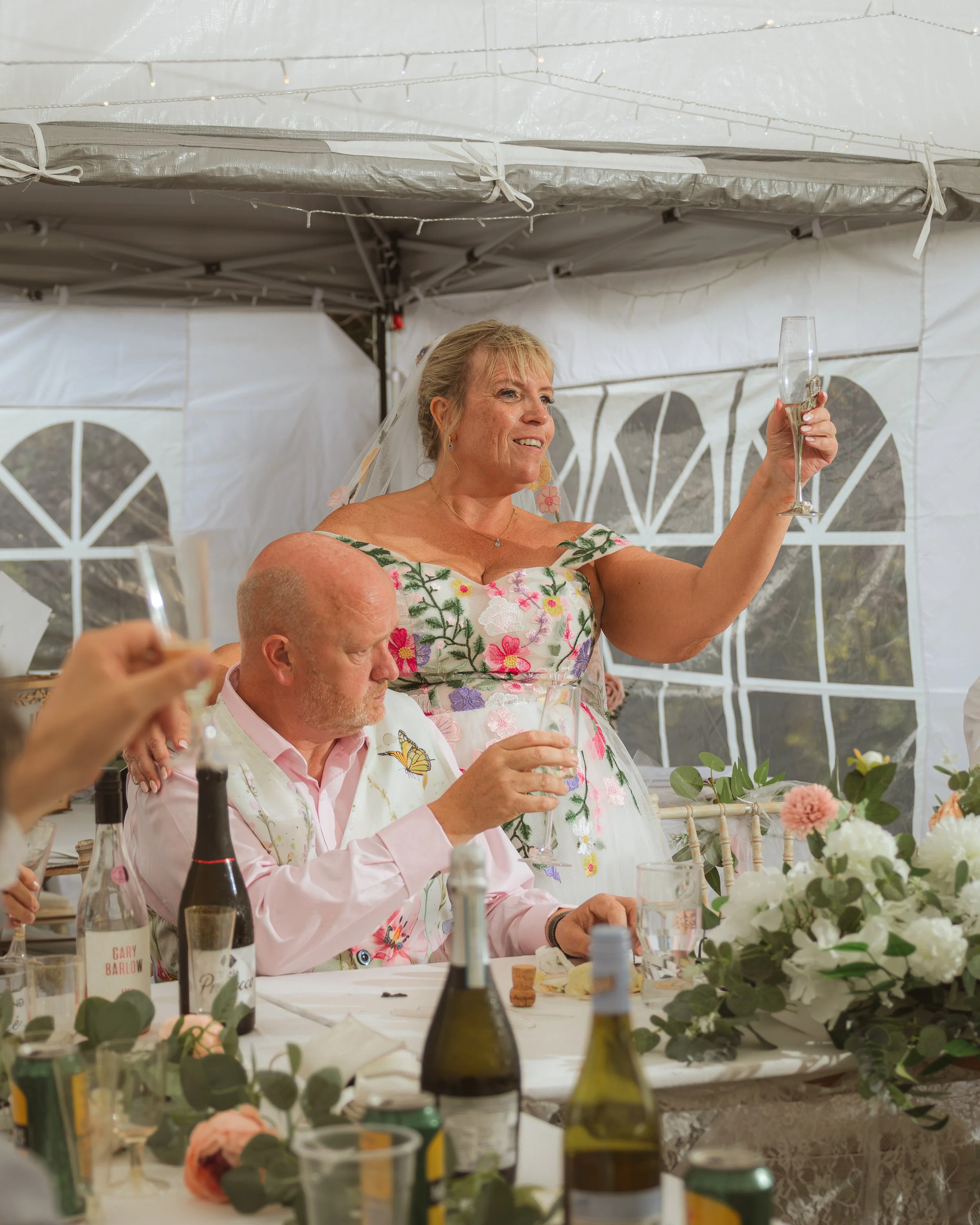 A woman in a floral dress and a veil holding a champagne flute at a celebration. She is smiling and looks happy. There are two bottles of wine and a floral centerpiece on the table.