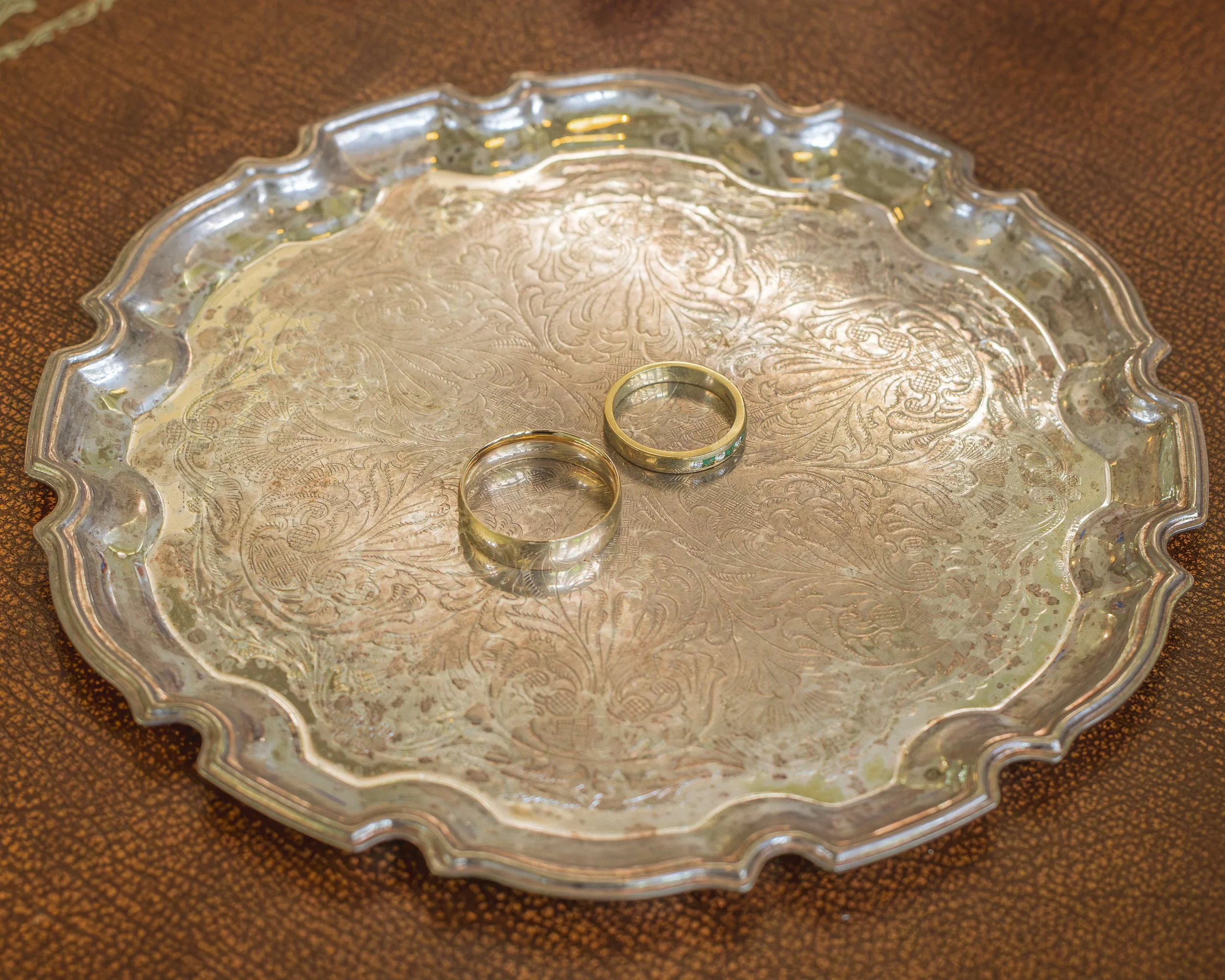 A silver tray with intricate engraved patterns and scalloped edges, resting on a textured brown surface, with two rings placed on it.