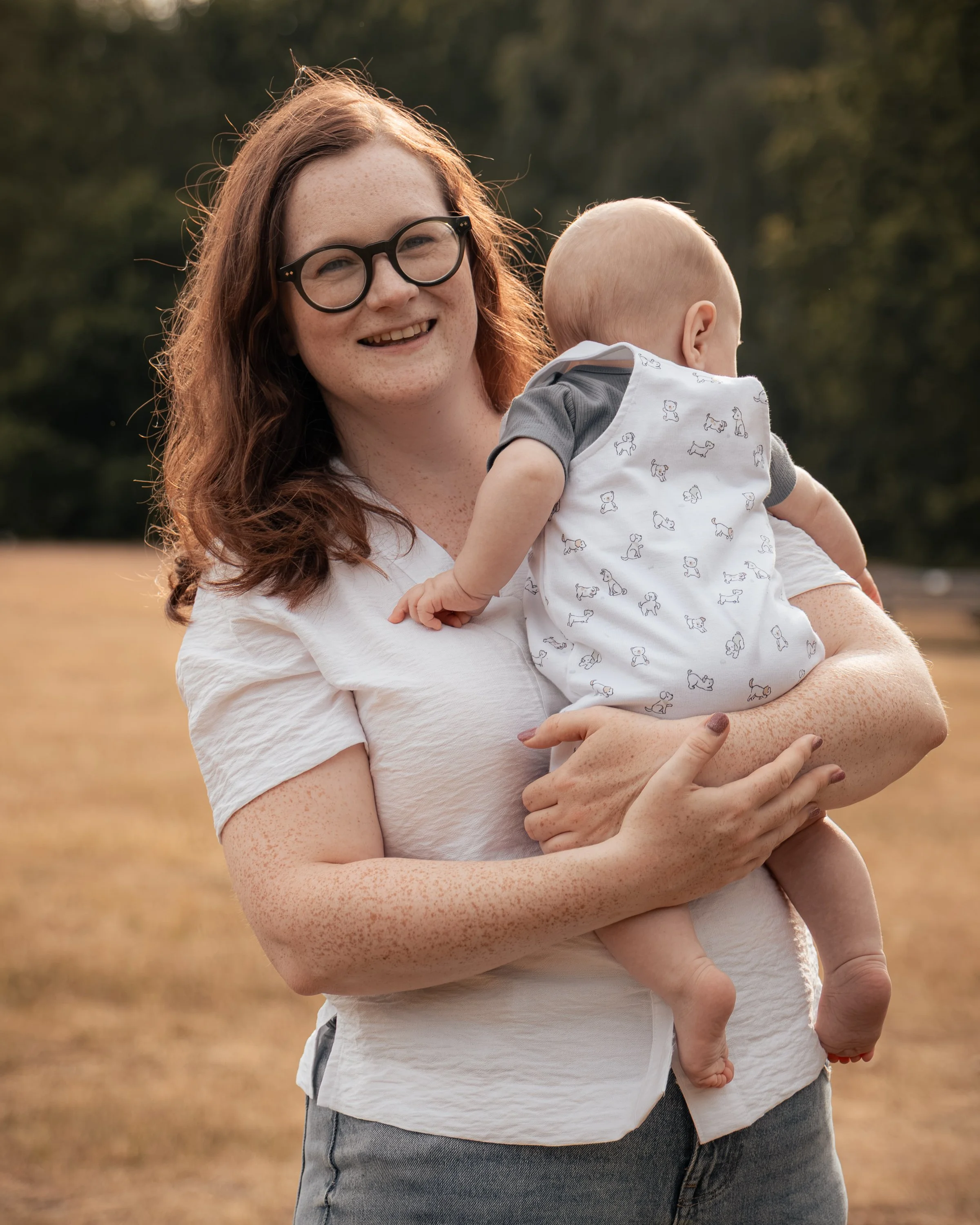 A woman with curly red hair, glasses, and light skin, smiling while holding a baby in an outdoor setting during golden hour.