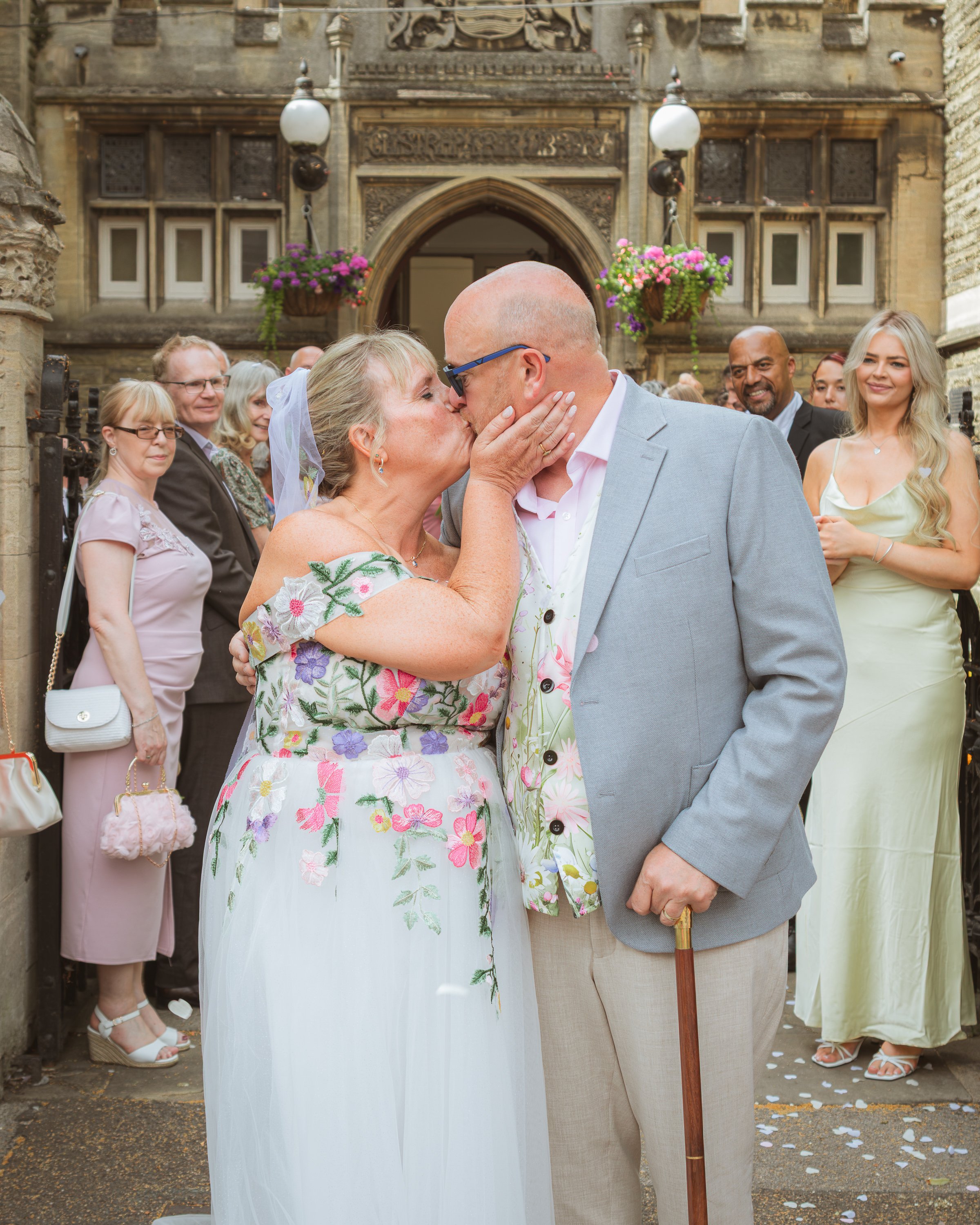 A wedding celebration outdoors with a couple kissing, surrounded by guests. The woman wears a floral dress and a veil, and the man sports a light gray suit and holds a cane. The background features a stone building with hanging flower baskets and vin