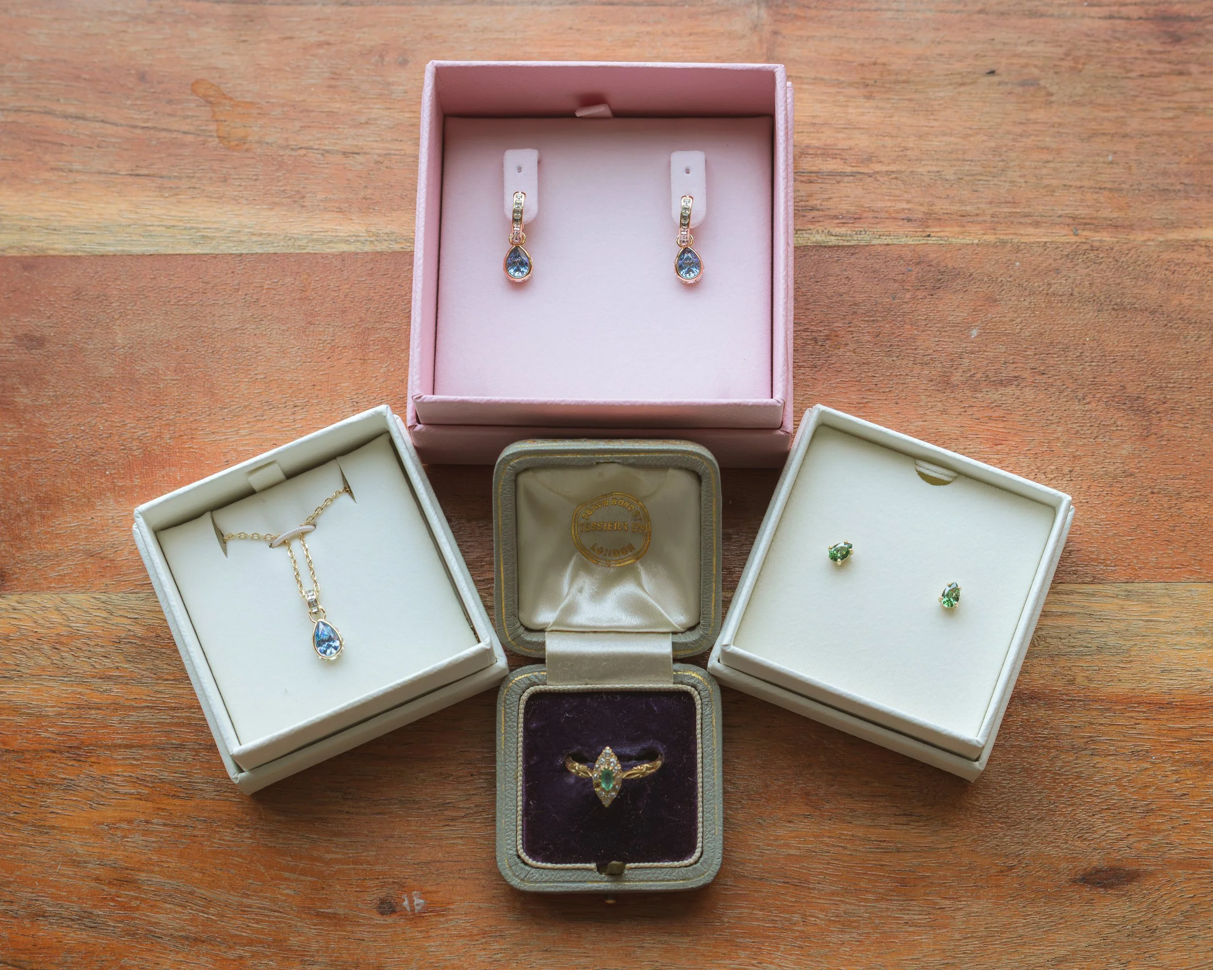 A pink jewelry box containing a pair of earrings with blue gemstones, and a white open jewelry box with a gold necklace, alongside a gray box with a purple lining, all placed on a wooden surface.