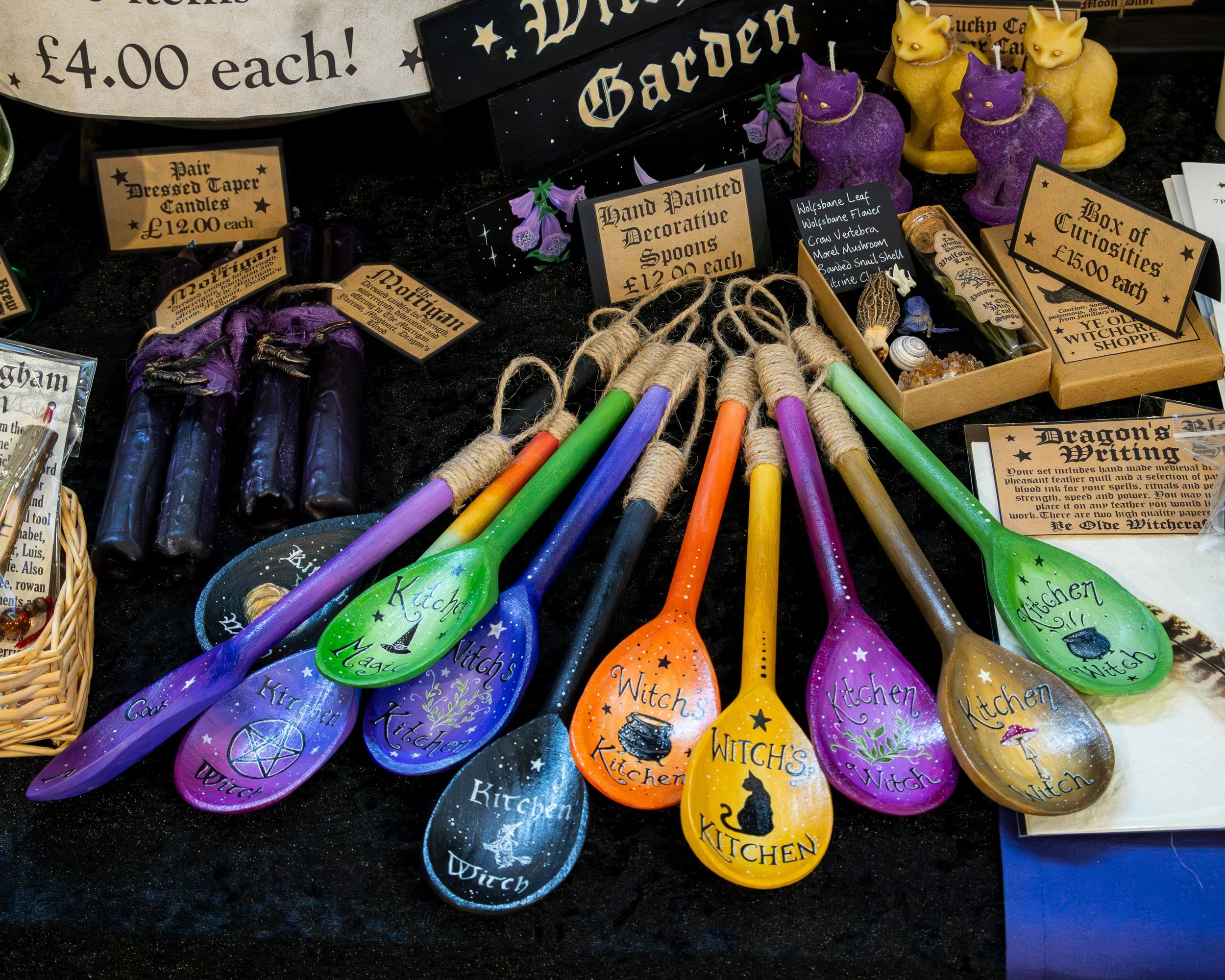 Colorful decorative spoons and candles with signs indicating prices, surrounded by various Halloween-themed items like black candles and witchcraft symbols, at a market stall.