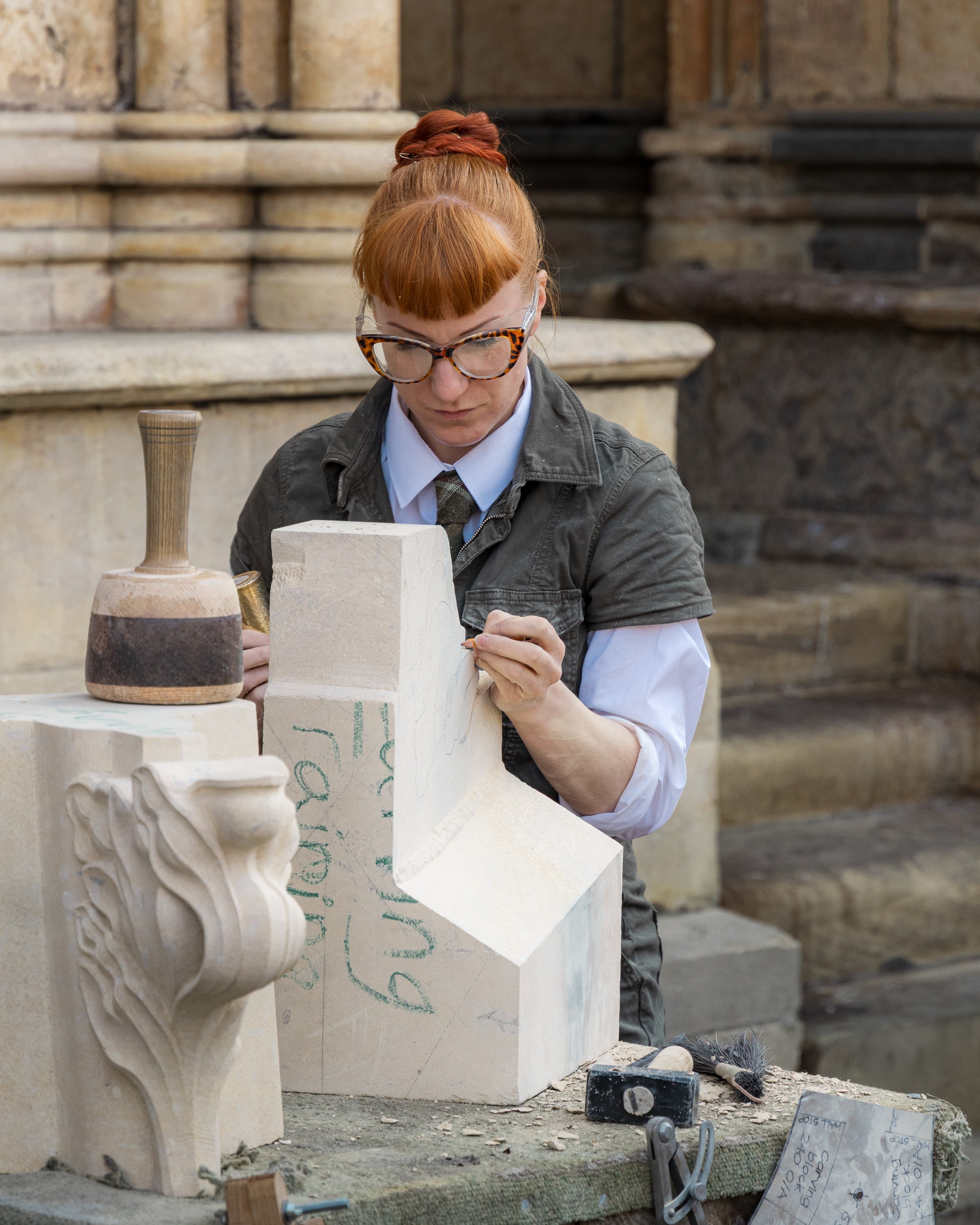 A woman with red hair, glasses, and a gray jacket working on a stone sculpture of a face in an outdoor setting with stone steps in the background.