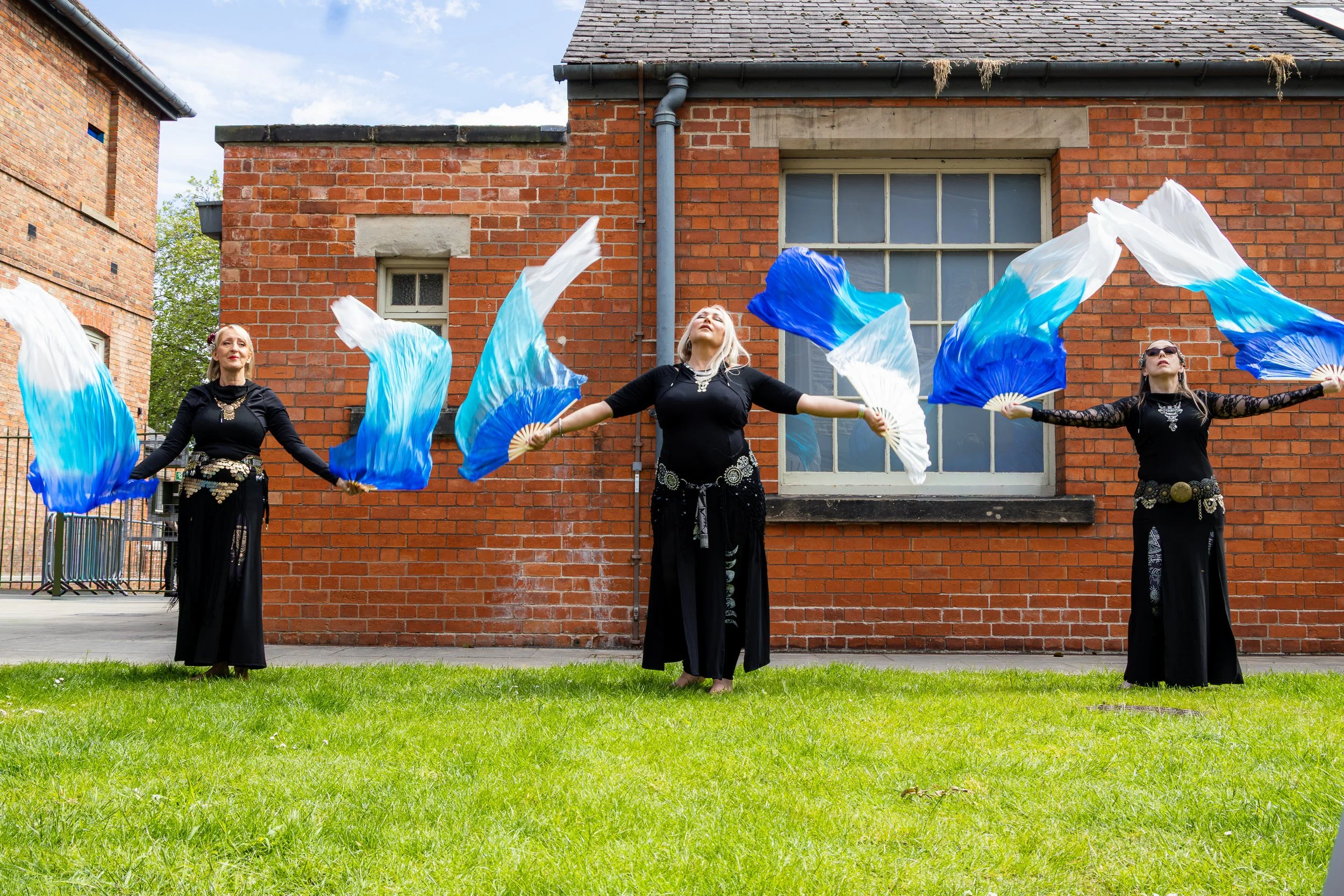 Three women dressed in black performing a dance with large blue and white fans in an outdoor courtyard with a red brick building in the background.