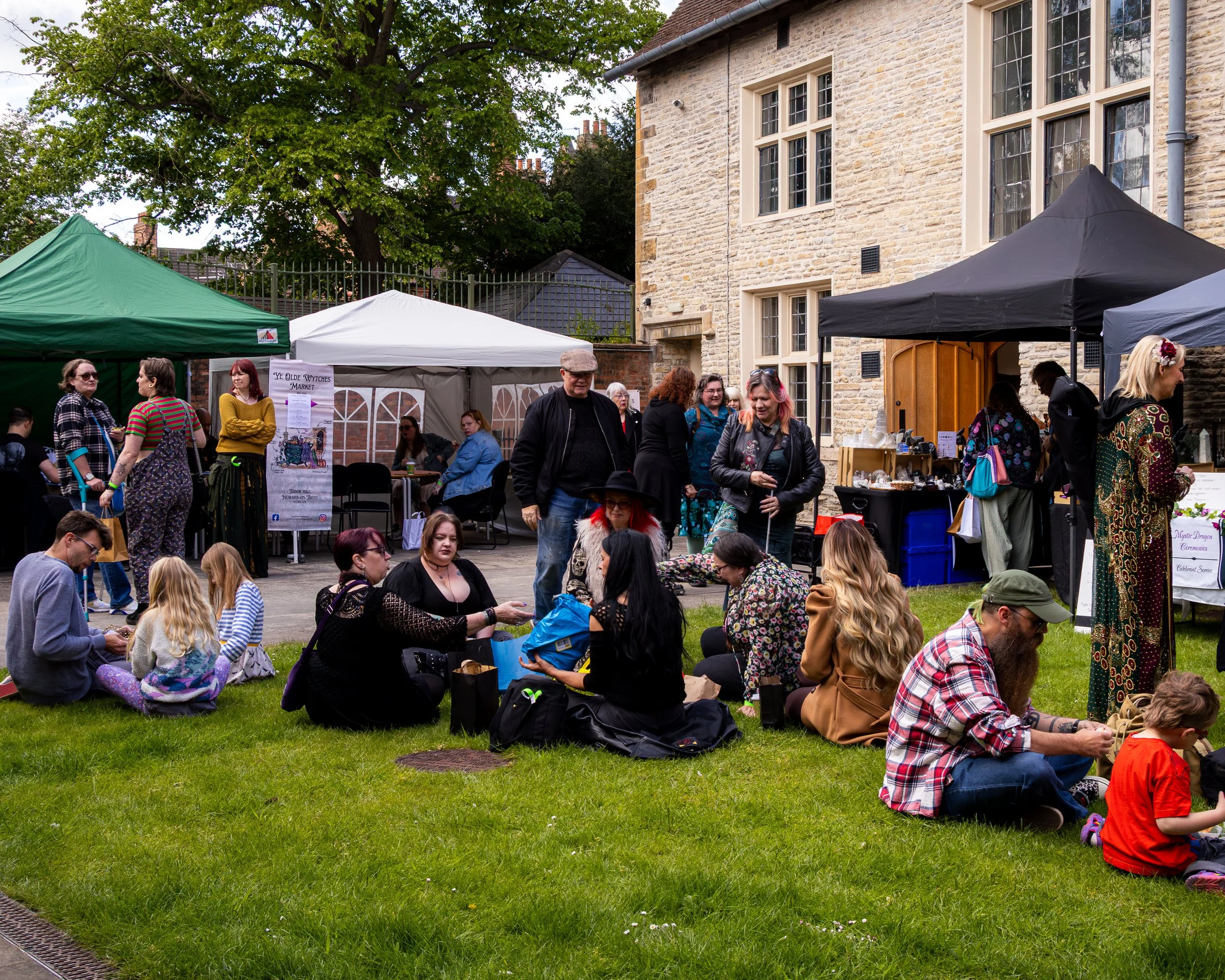 People attending an outdoor market event in front of a stone building with large windows, green grass and trees, and vendor tents in various colors including green, white, and black.