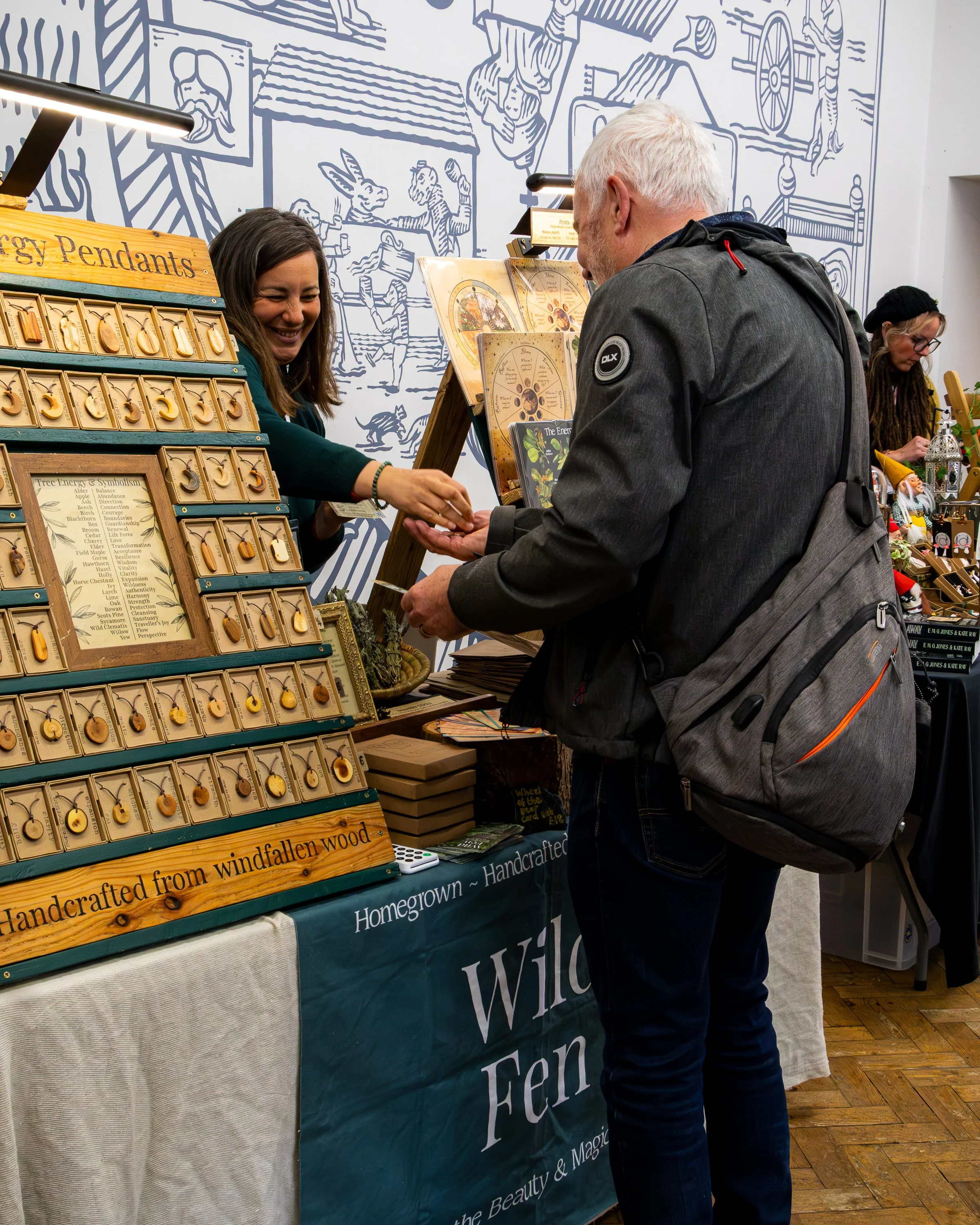 A woman smiling and handing a small object to a man at a booth with wooden sign reading 'Energy Pendants' and displays of various pendants on a board. The background features a mural with cartoon-like drawings of animals and people.