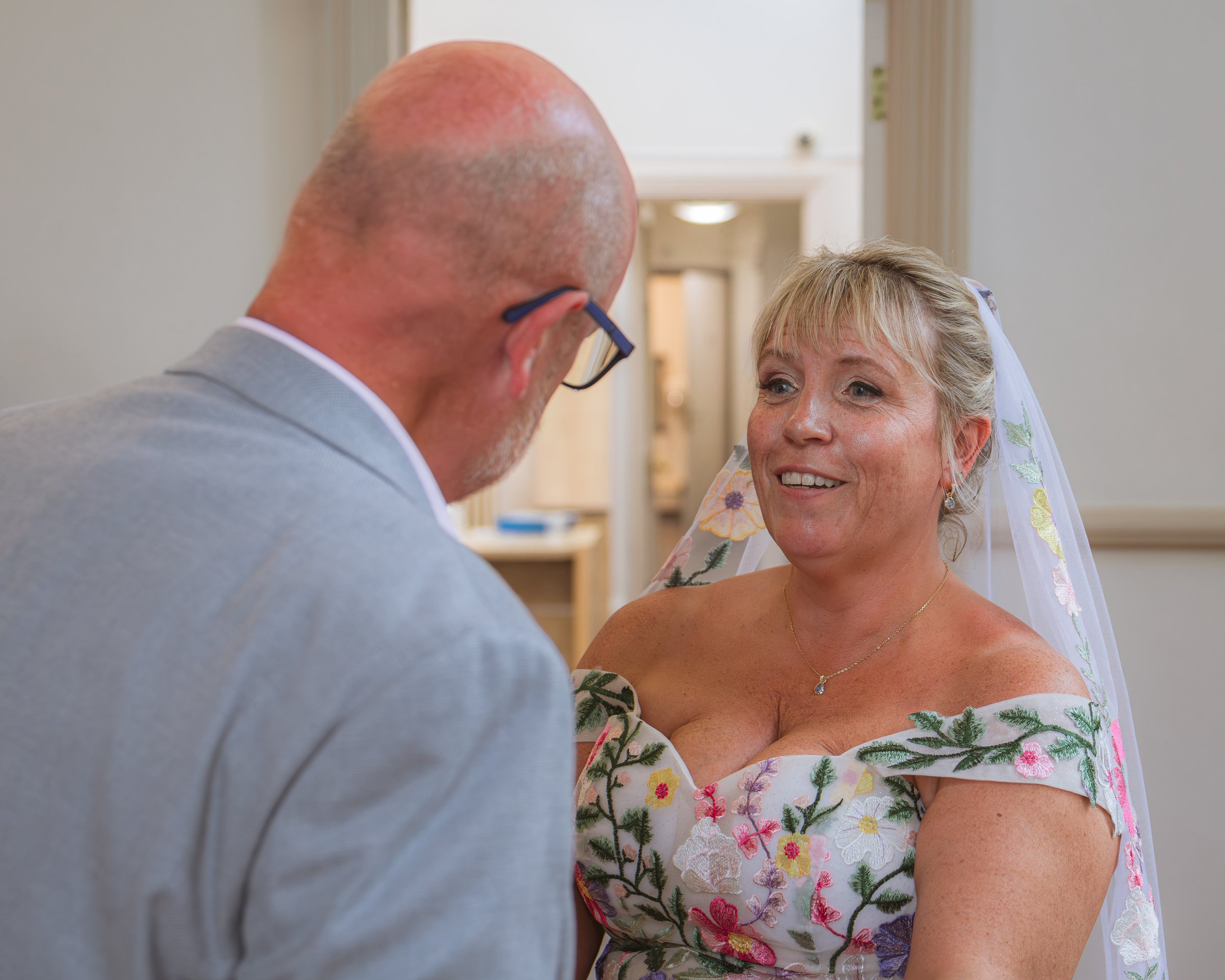 A bride with short blonde hair and a floral dress, smiling and looking at an older man, possibly at her wedding.