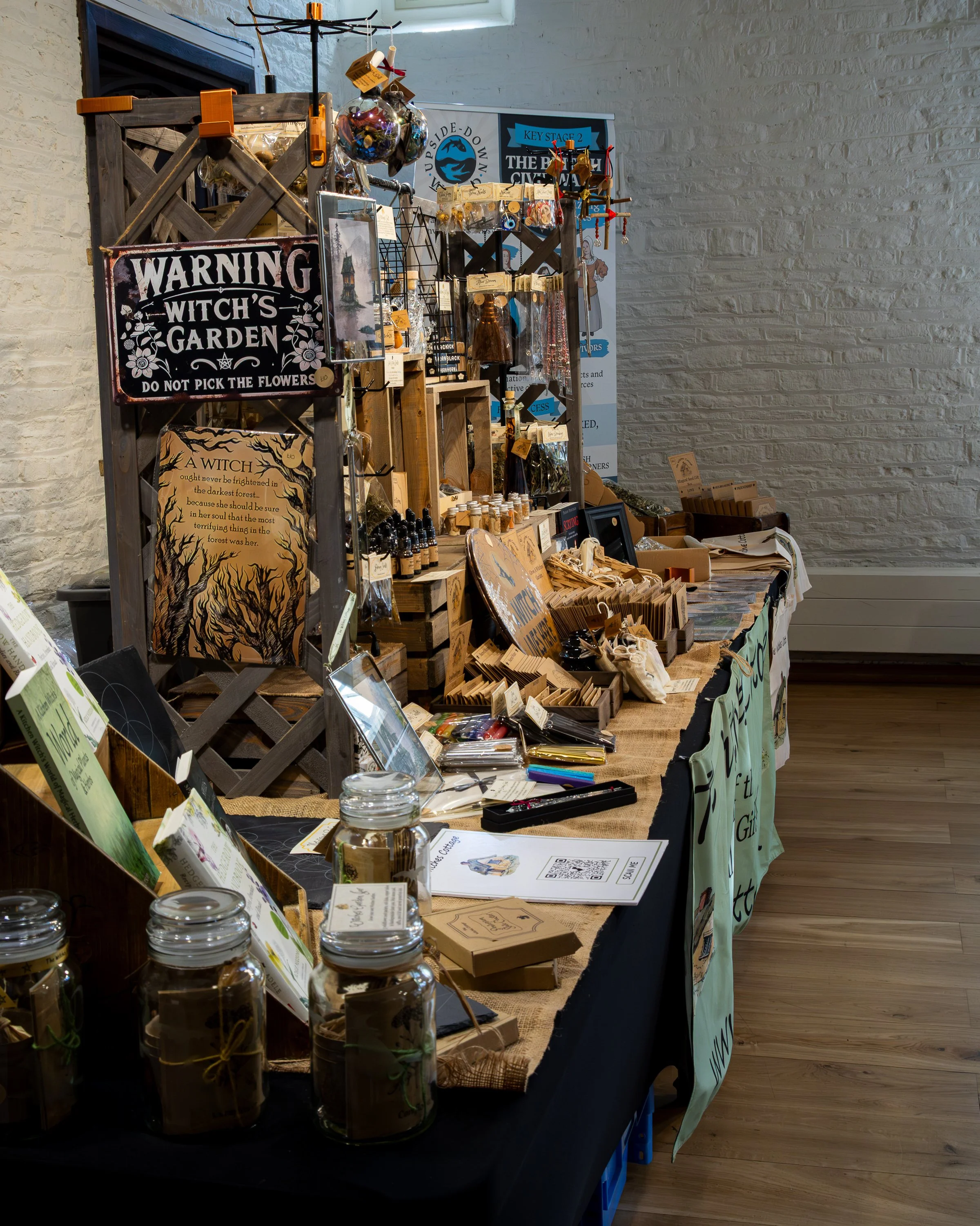 A market stall displaying various products, including jars, postcards, jewelry, and decorative items, set against a white brick wall with signage in the background.