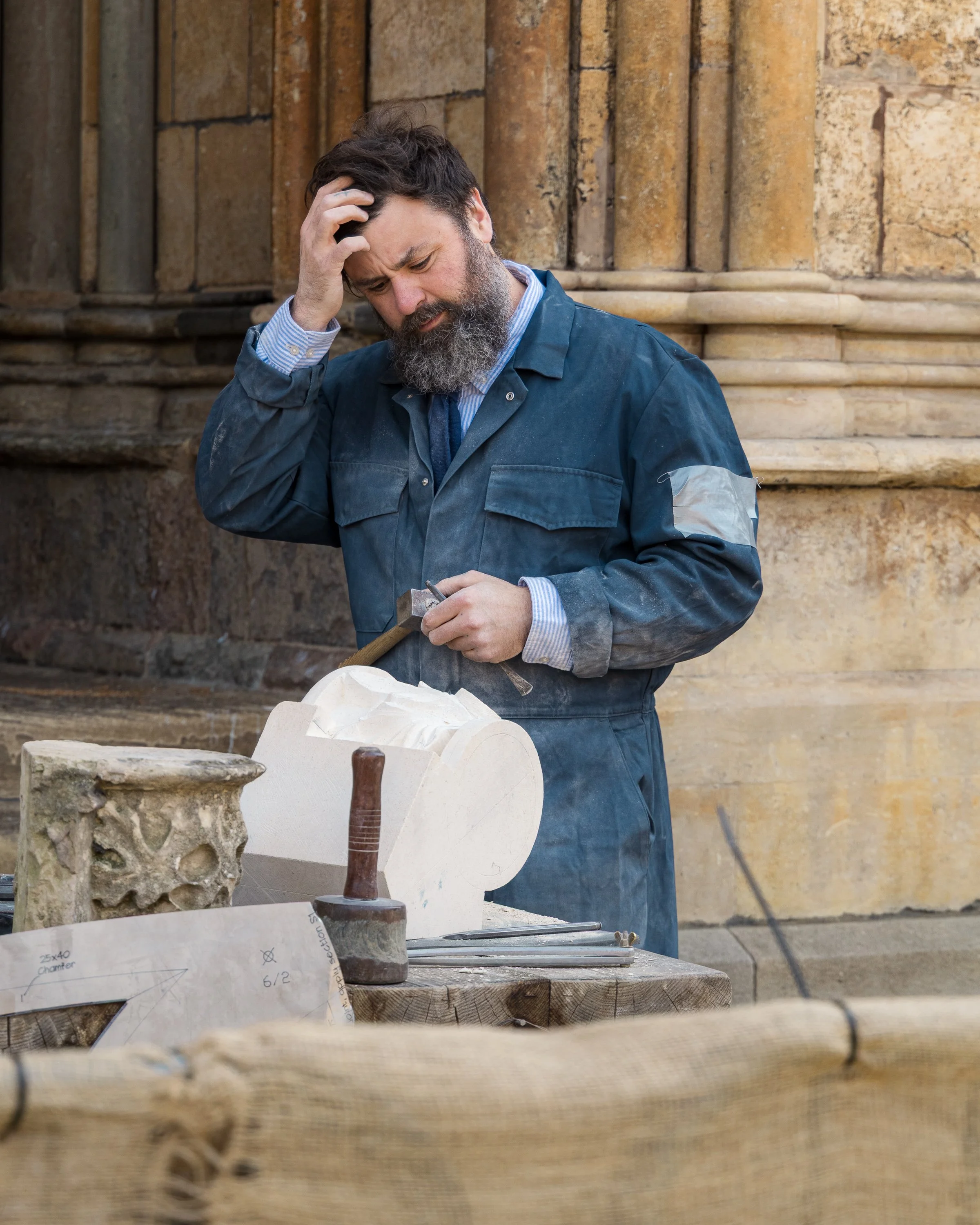 A man with a beard, wearing a blue work jacket, holding a hammer, standing outdoors against an ancient stone building, with woodworking tools and plans on a wooden workbench in front of him.