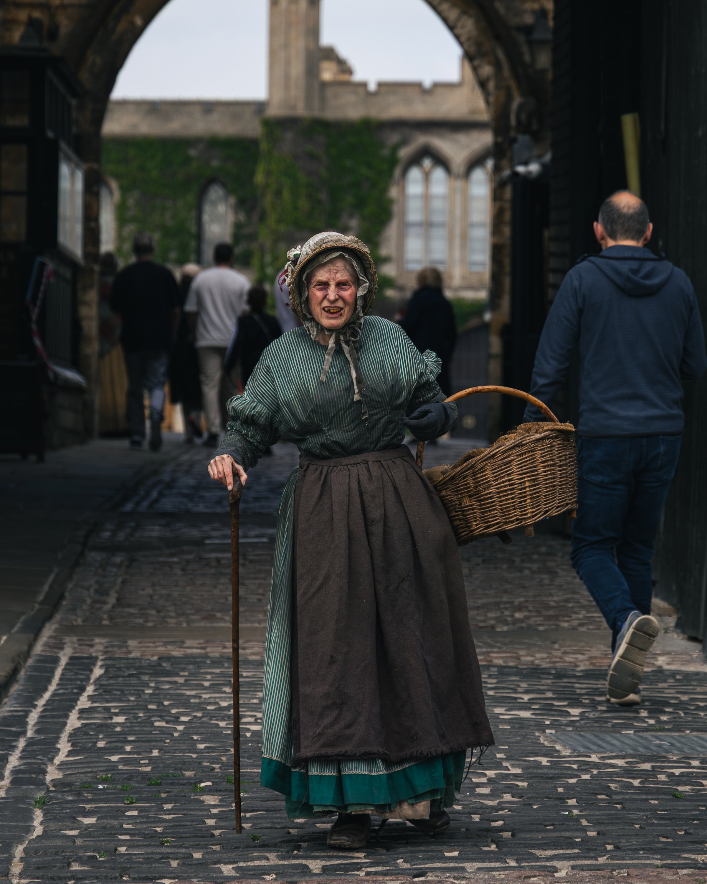 An elderly woman dressed in historical clothing, holding a cane and a basket, walking through a cobblestone street near a castle with gothic windows.