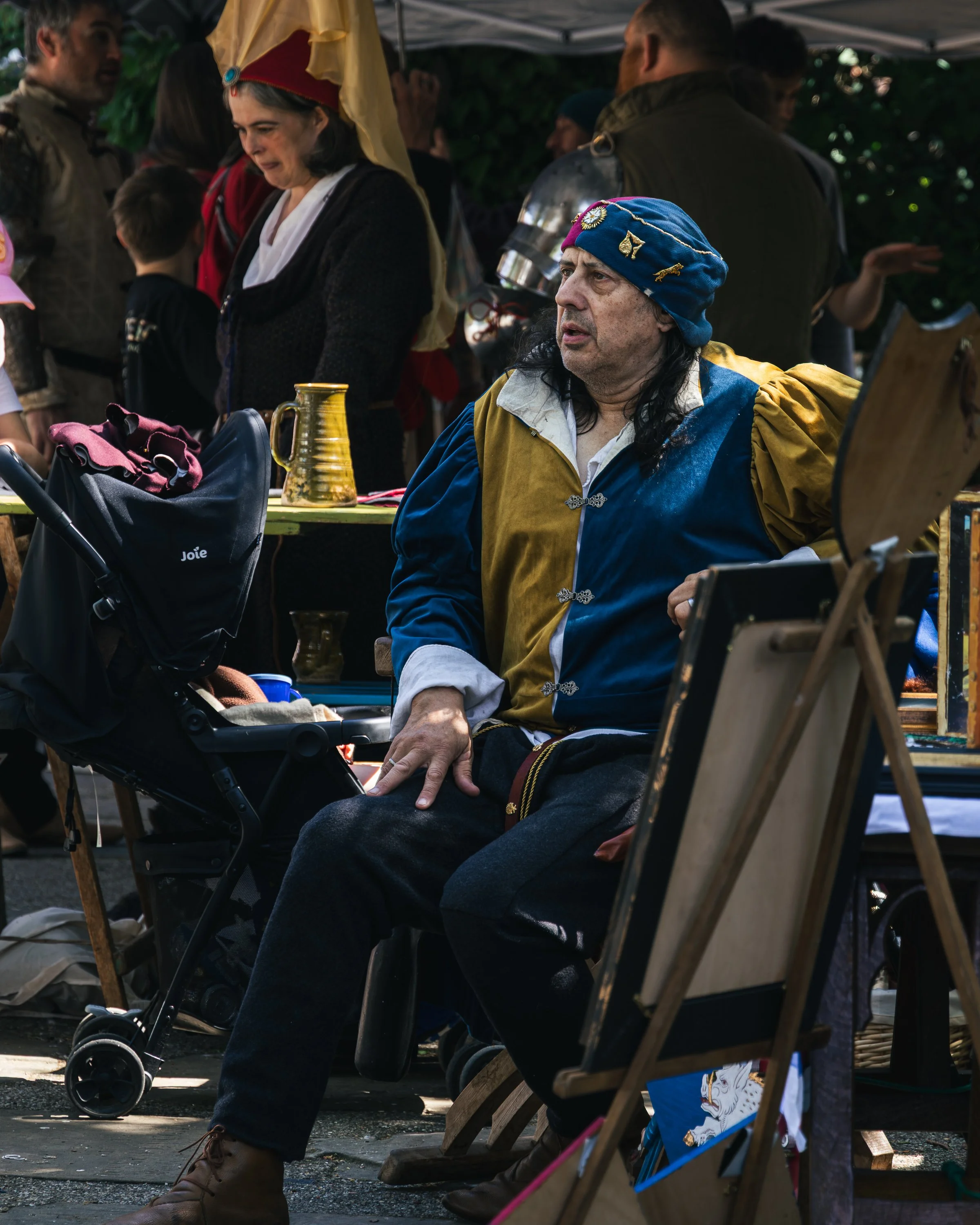 A man dressed in medieval or renaissance-style costume with a blue cap decorated with various embroidered symbols and a yellow and blue tunic, sitting on a wooden bench at an outdoor event. The background includes other people, some in costumes, and 