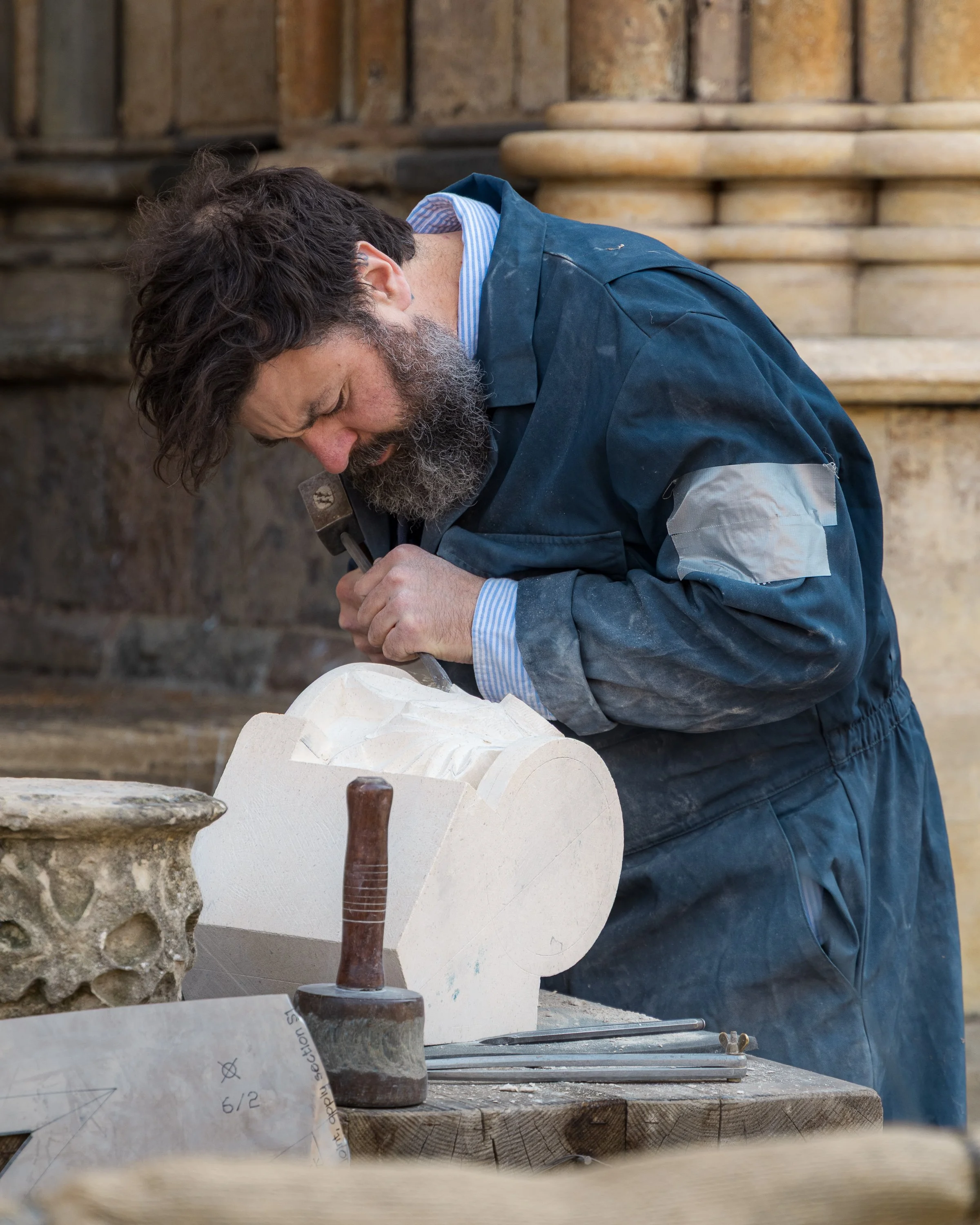 A man with dark hair and a beard is working on a carving project at a workbench, with stone statues and tools visible around him.