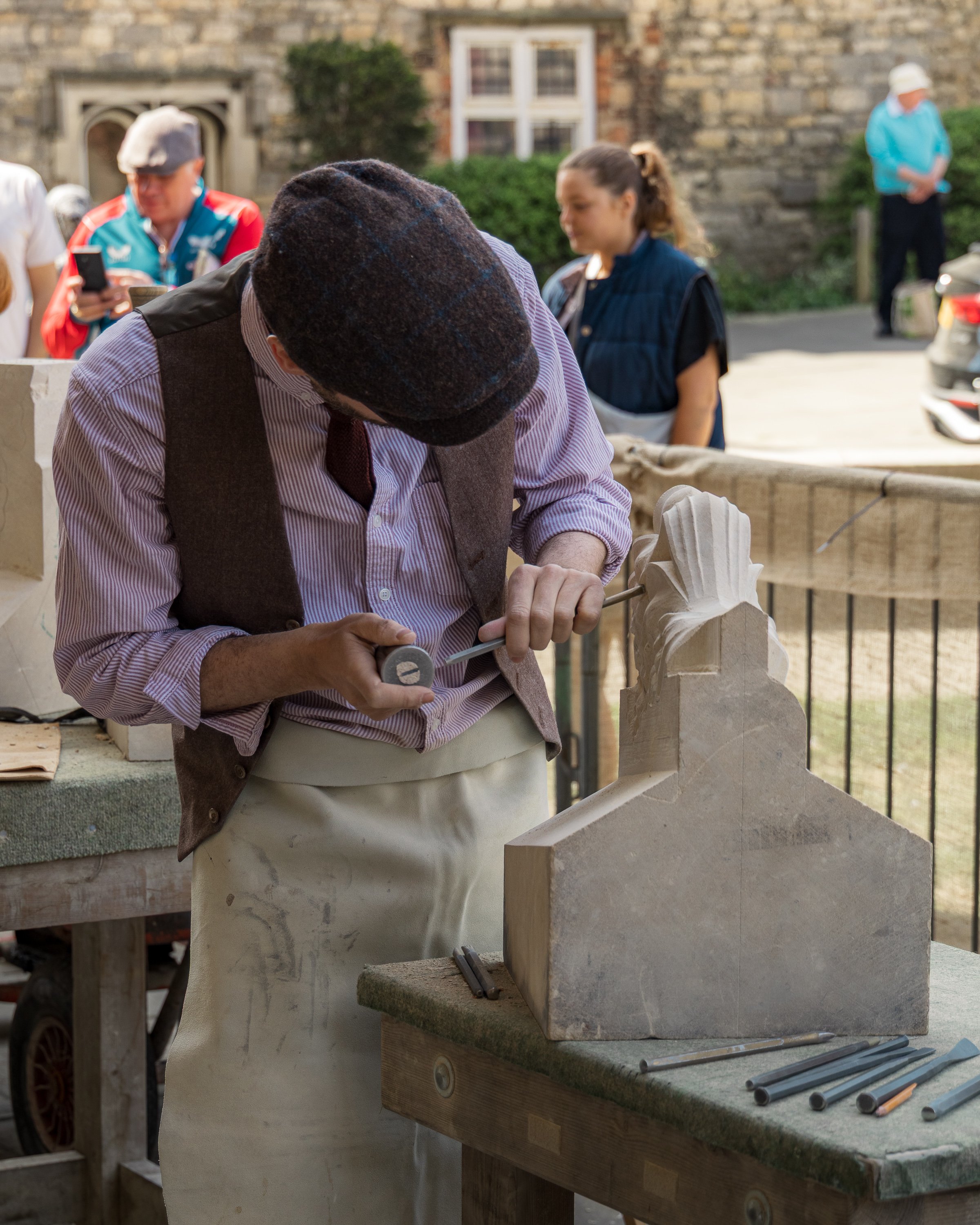 A craftsman carving a sculpture out of light-colored stone on a workbench outdoors. Several metal tools are laid out on the bench. In the background, a woman and a man are seen; the man appears to be photographing or looking at his phone, and the wom
