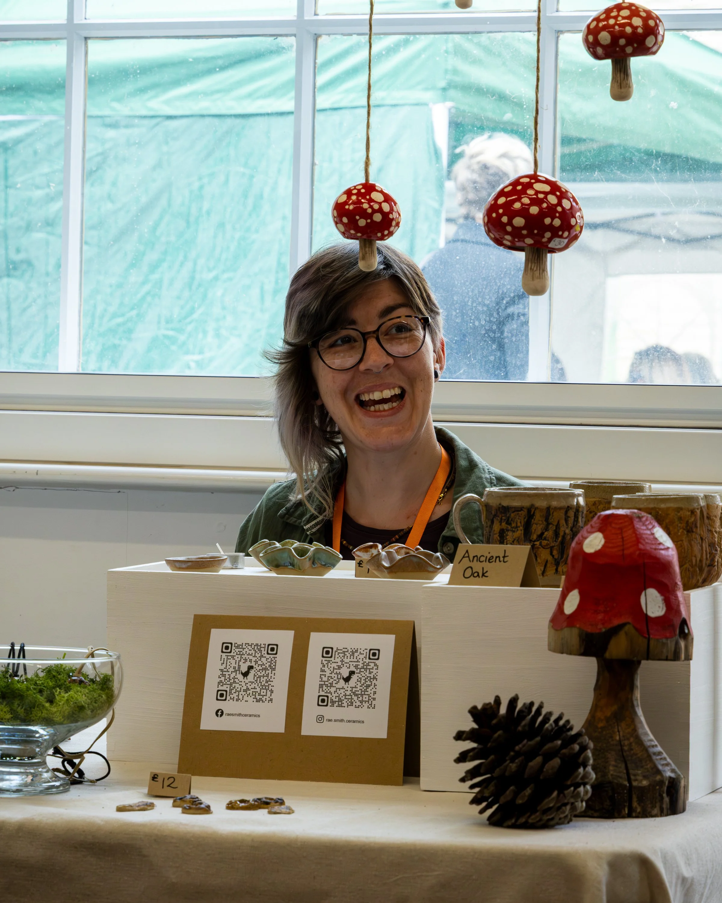 Display of ceramic items at an indoor market or craft fair. Items include small bowls, mushroom-shaped decorations hanging from the ceiling, a map, a pine cone, and a wooden figure with a red mushroom cap. A woman with glasses is smiling behind the t