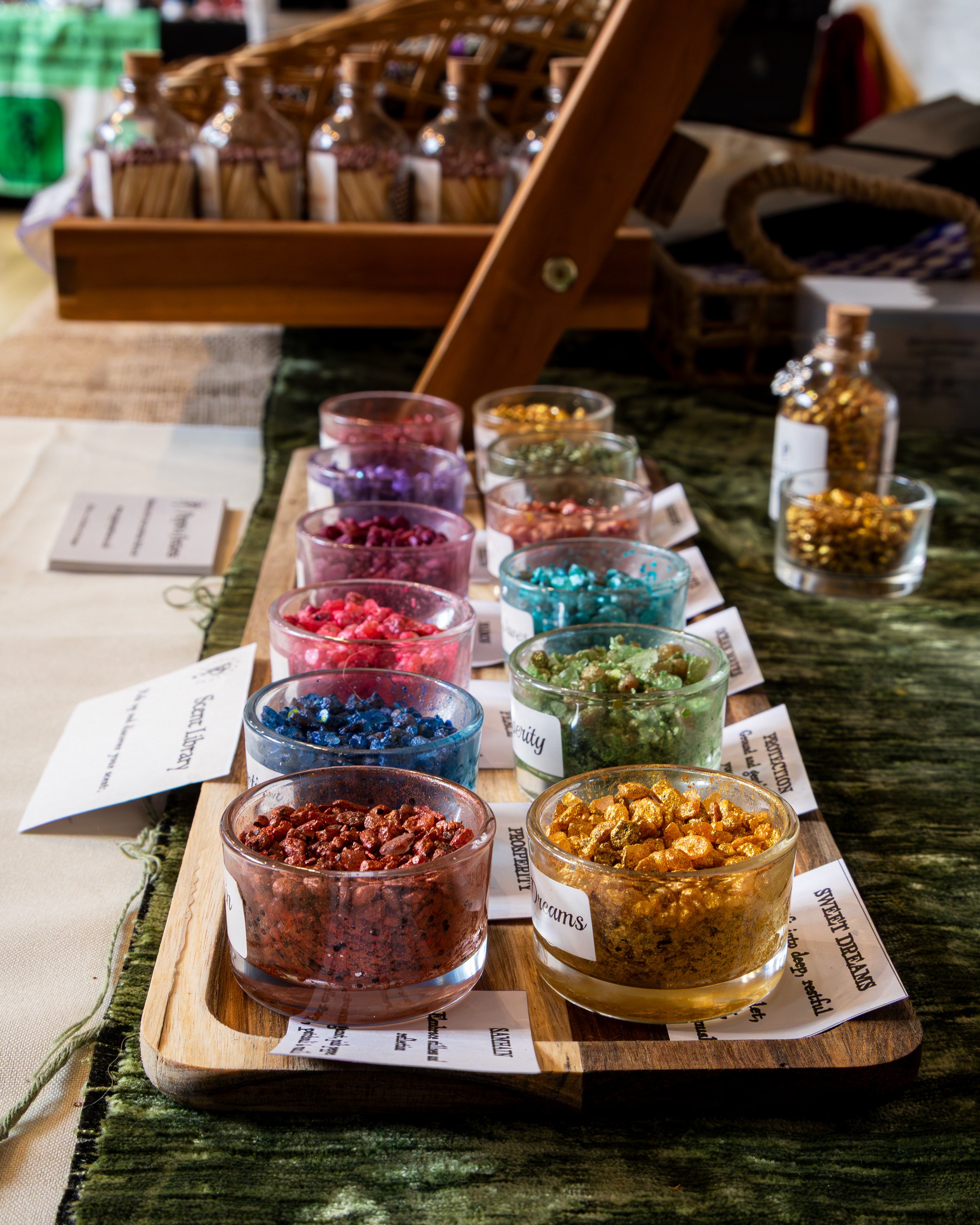 A variety of colorful crushed gemstones in small glass bowls arranged on a wooden tray, each labeled with different shades and types of stones for display.