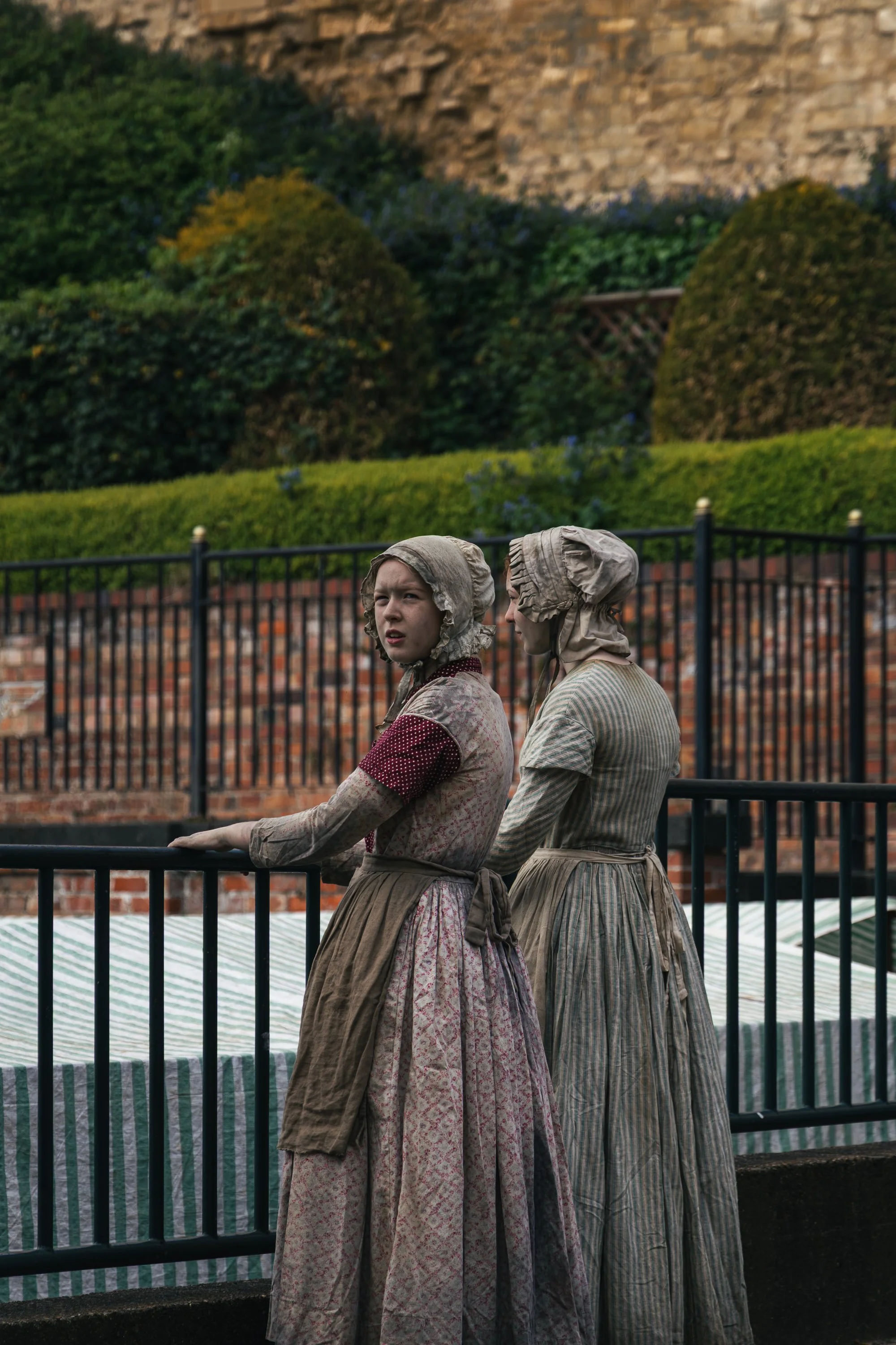 Two women dressed in 19th-century colonial attire with bonnets standing behind a metal fence, with a brick and stone wall, greenery, and trees in the background.