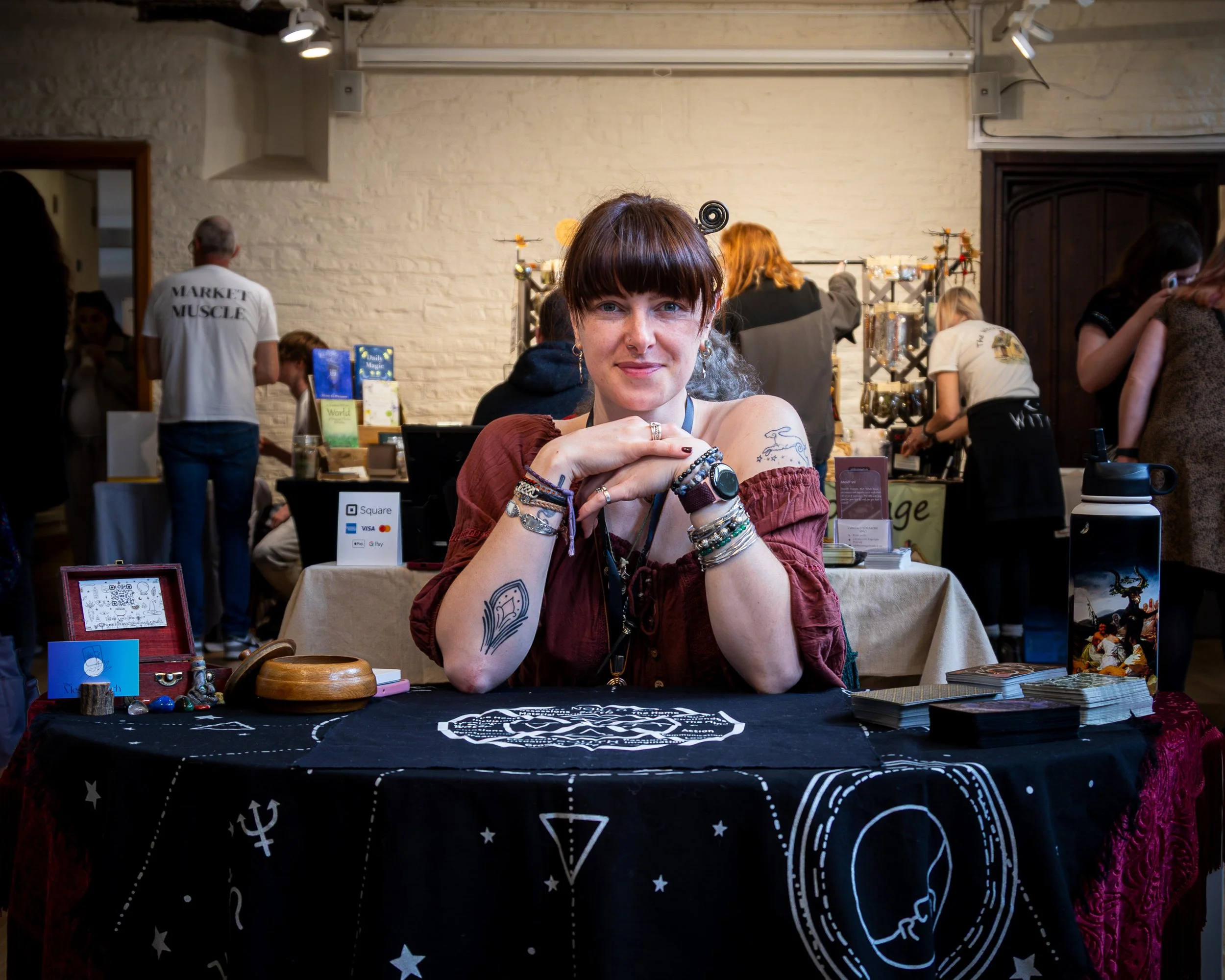 A woman with tattoos and wrist jewelry sitting at a table with tarot and oracle cards, jewelry, and decorative objects, smiling at the camera inside a busy marketplace or craft fair with people, booths, and displays in the background.