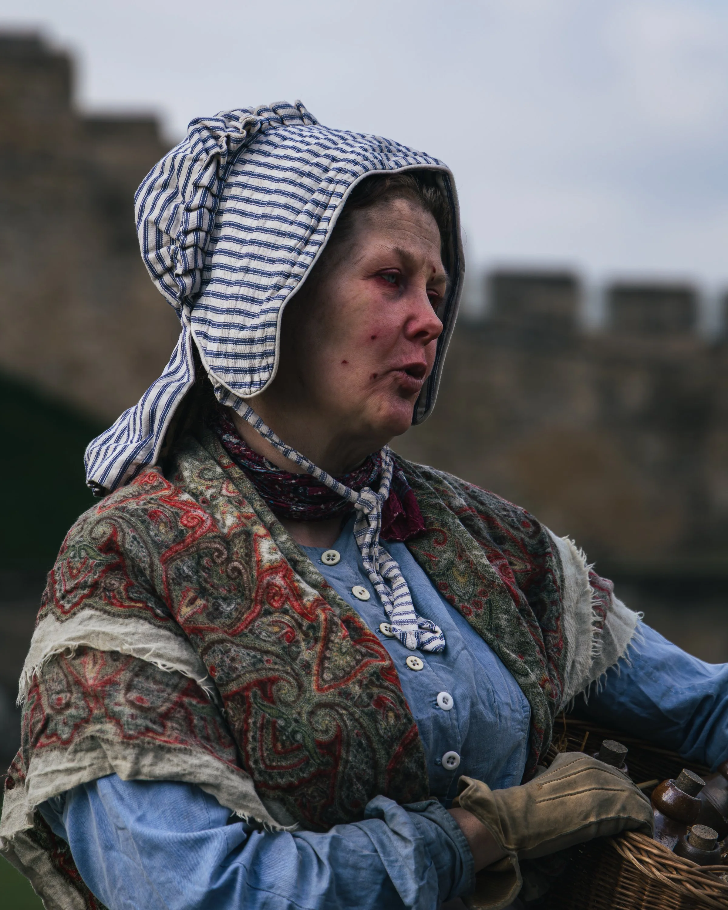 A person with a weathered face, wearing a striped cloth hood and a patterned jacket, appears to be outdoors on a cloudy day. They are holding a woven basket with bottles inside, and possibly wearing work gloves.