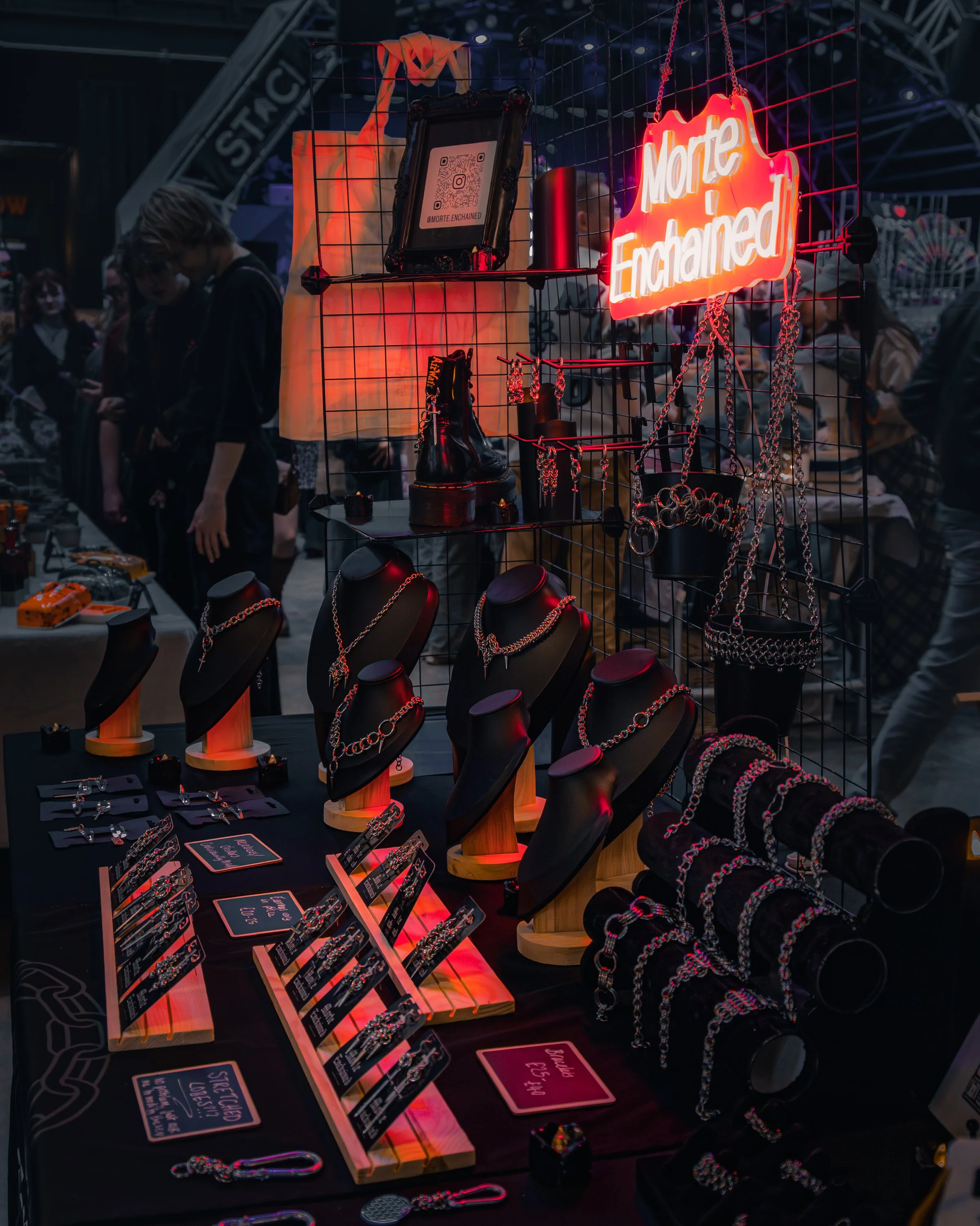 A display of jewelry including necklaces, rings, and other accessories on black mannequin busts and a table, illuminated with red neon lighting. There is a bright neon sign that reads 'More Enchained' on a wire grid, with other decorative items like 