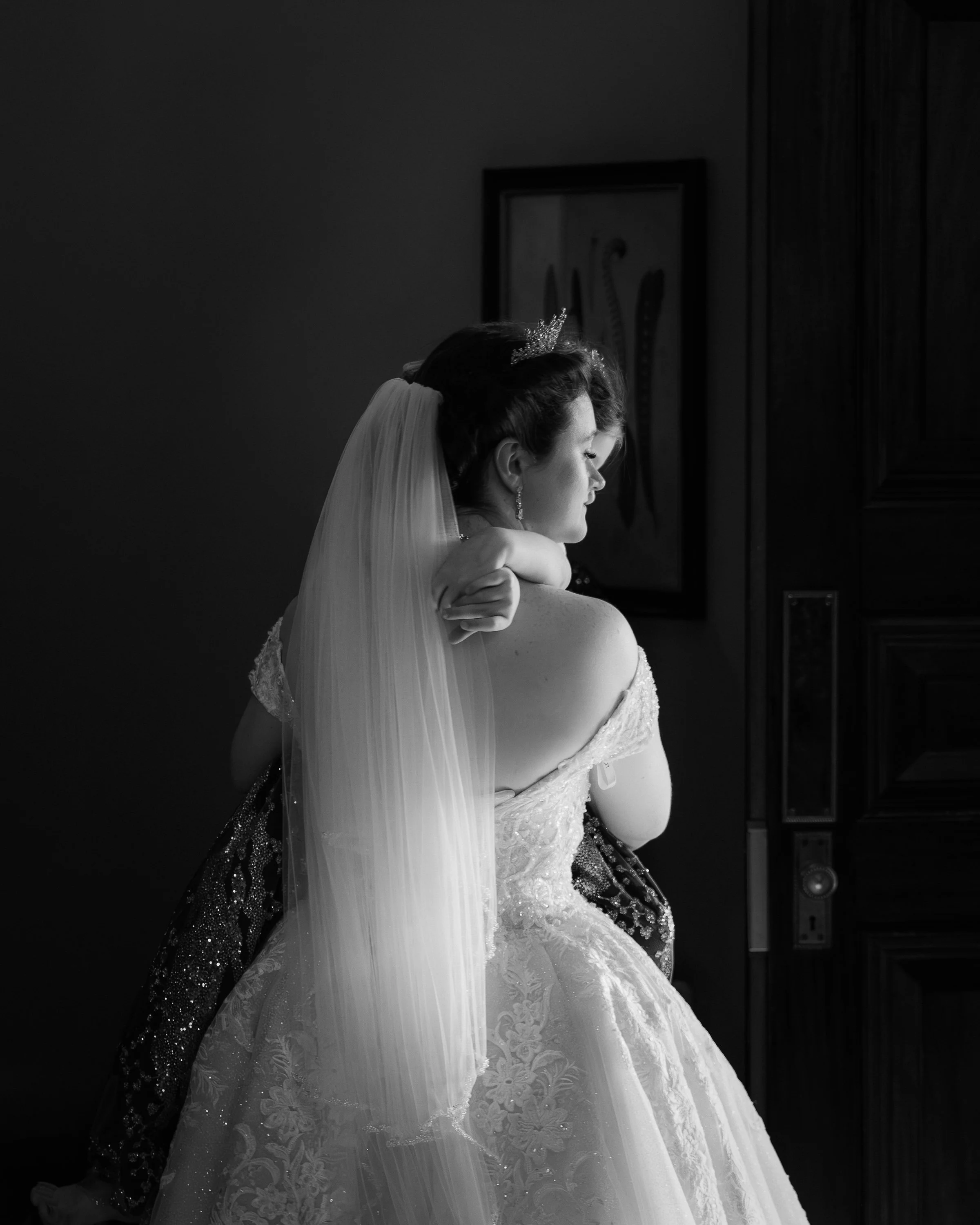 Black and white photo of a bride with her eyes closed, wearing a strapless wedding gown, a veil, and a tiara, standing with her arm crossed over her shoulder in a dimly lit room.