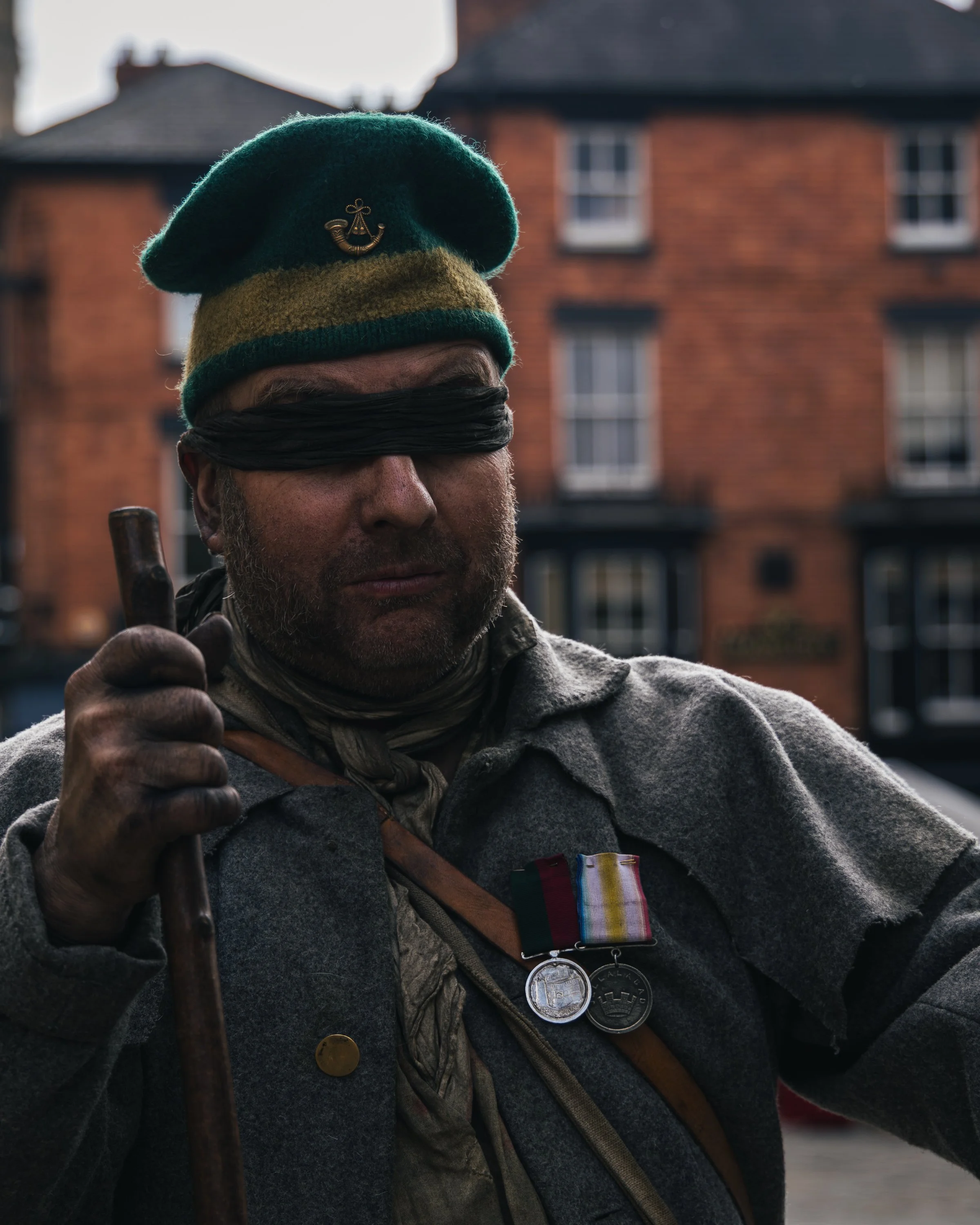 A man wearing a green beret with a badge, blindfolded with a black cloth, holding a wooden staff, dressed in a gray jacket with medals and a ribbon, standing outdoors in front of brick buildings.